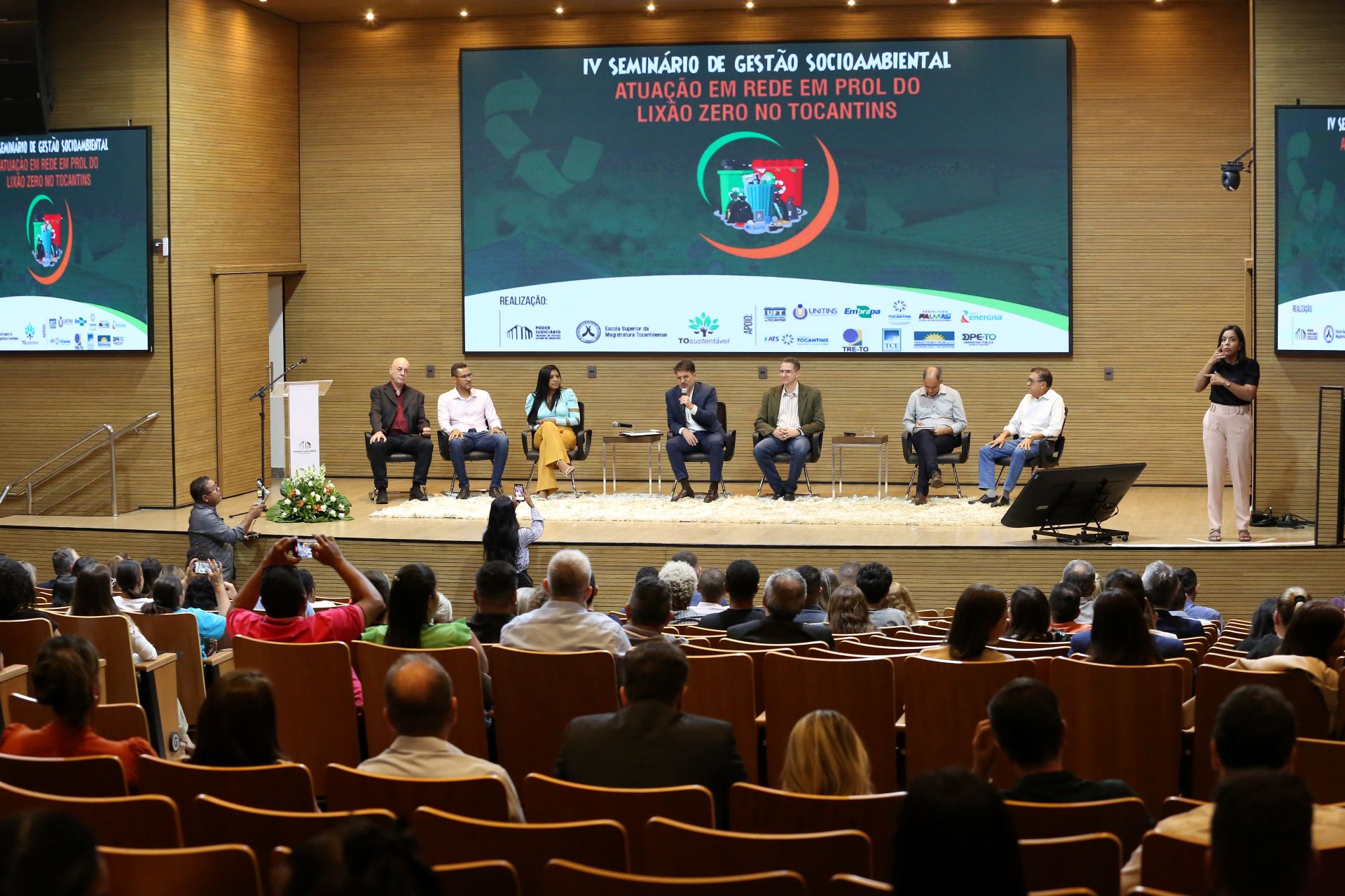 Imagem mostra o auditório cheio de cadeiras e pessoas sentadas à frente uma equipe discute sobre lixões