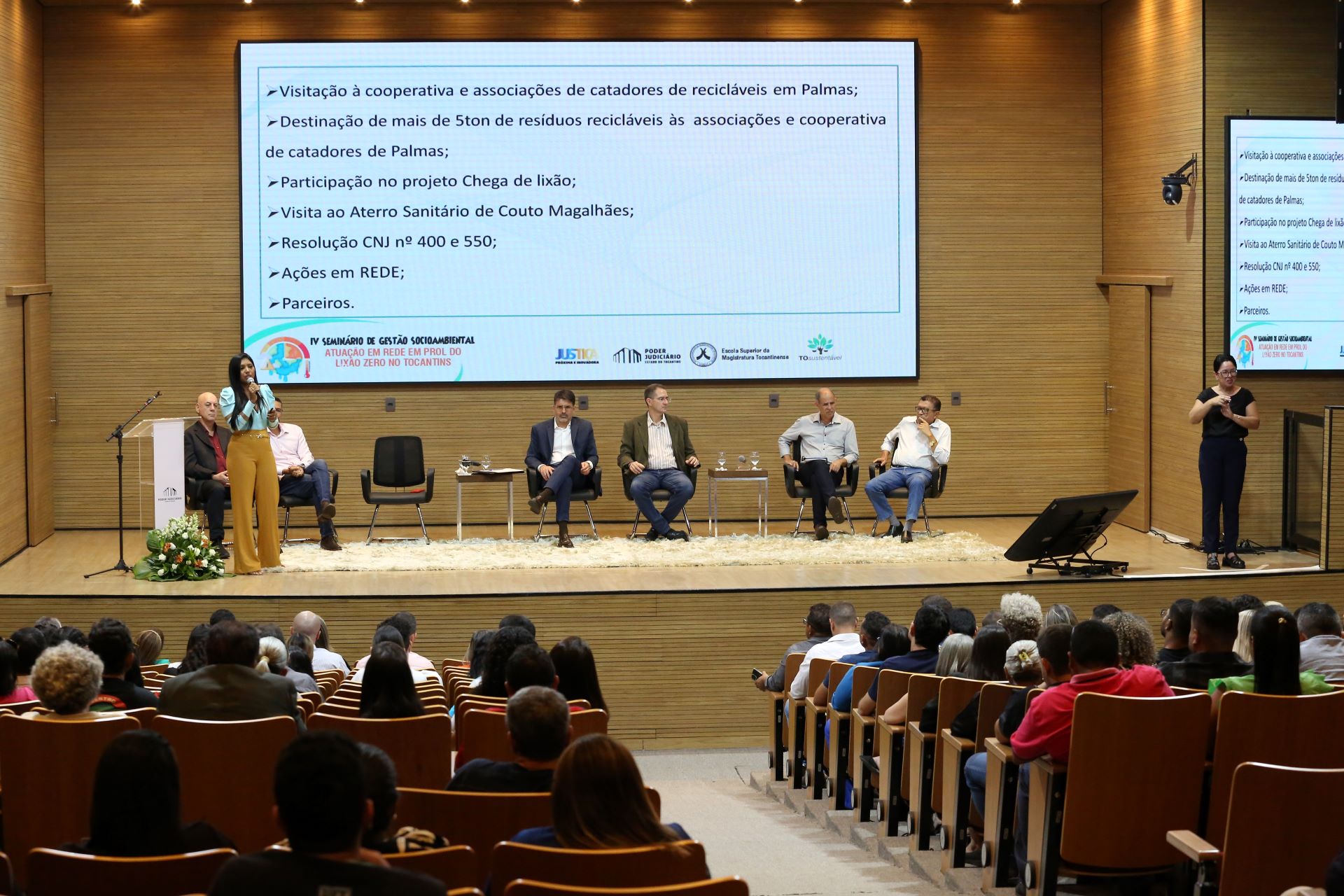 Uma mulher de verde e amarelo está em cima de um palco falando para uma plateia