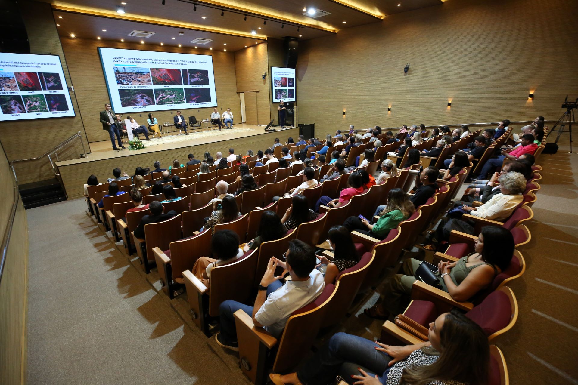 Imagem mostra o auditório cheio de cadeiras e pessoas sentadas à frente uma equipe discute sobre lixões