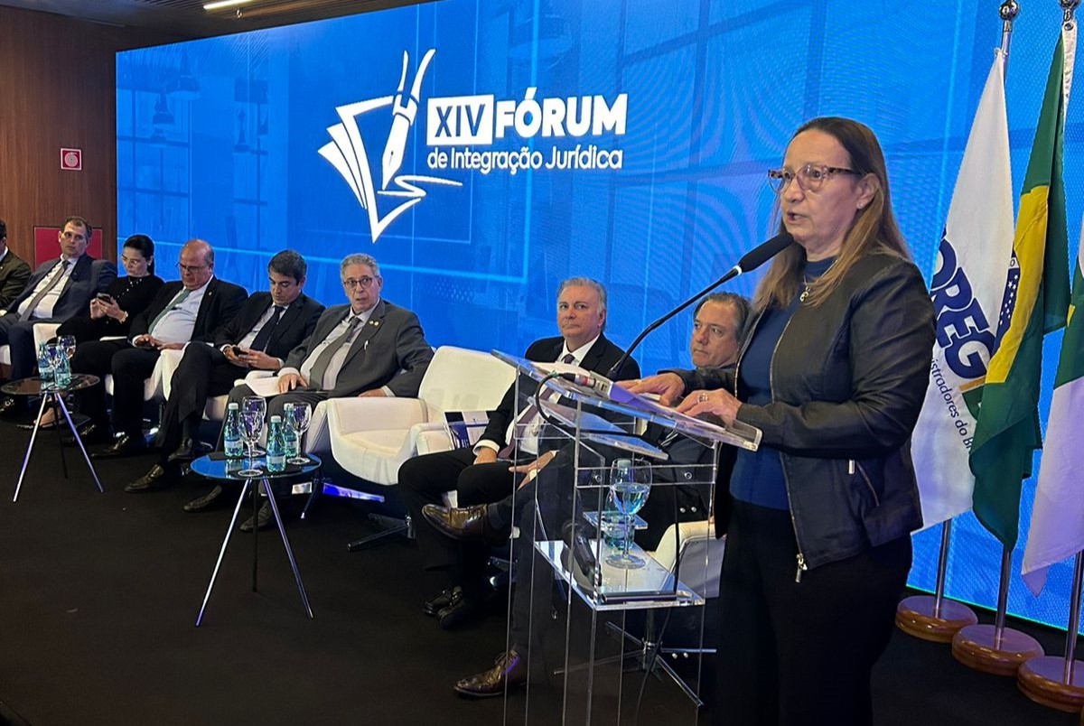 A imagem retrata uma mesa de discussão no "XIV FÓRUM de Integração Jurídica". O cenário é um palco com um pLano de fundo exibindo o nome e o logotipo do fórum. Sete pessoas estão sentadas em uma fileira no palco, todas vestindo trajes formais, e uma desembargadora está falando no púlpito com um microfone.