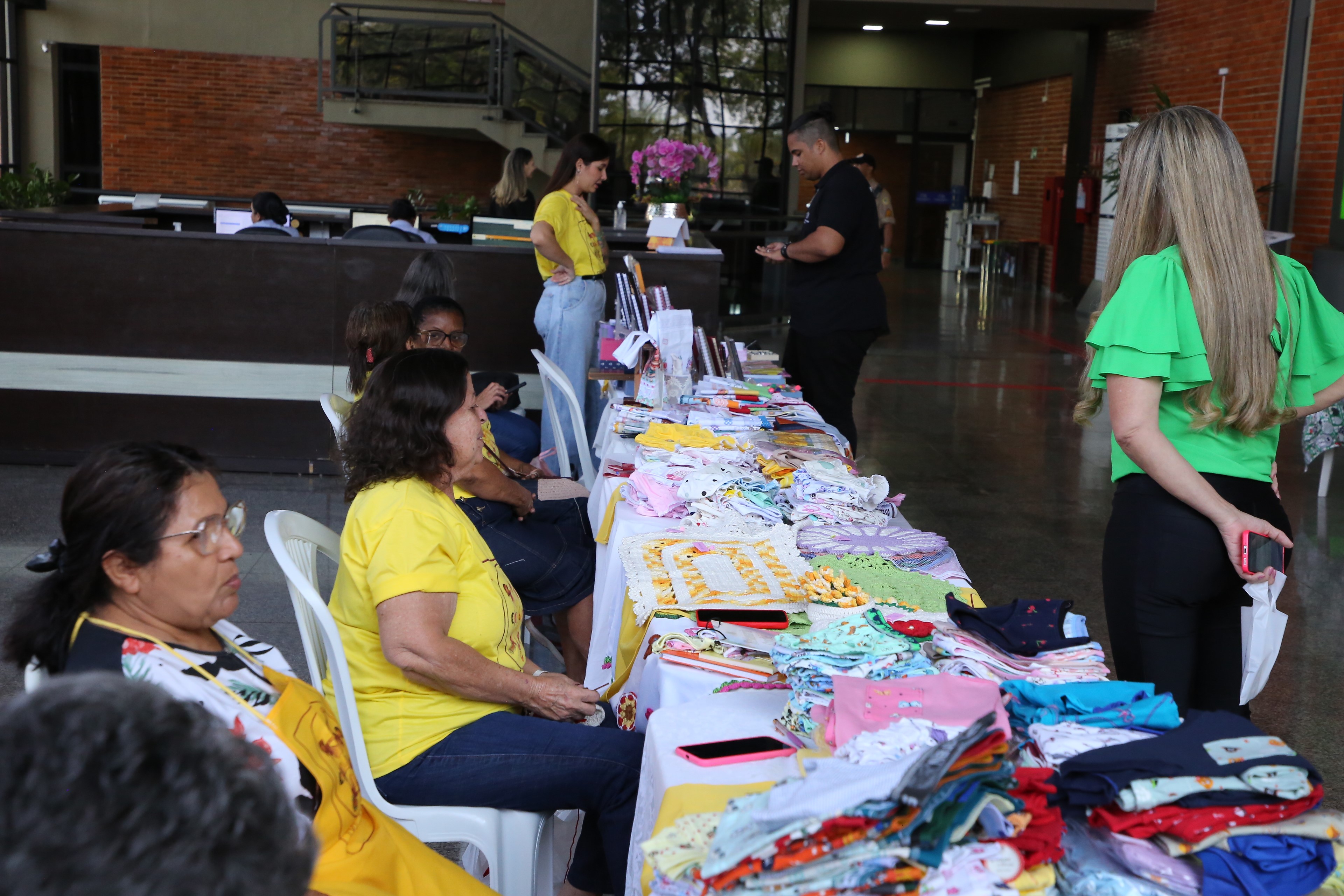 Fotografia colorida onde um homem de roupa preta e cabelo preso e uma mulher de cabelo loiro solto e camisa verde clara, e calça preta, estão de um lado de uma grande mesa, enquanto algumas mulheres de camisas amarelas e cabelos pretos estão do outro lado, conversando e apresentando produtos que estão dispostos em cima dessa longa mesa. Eles estão em um grande espaço com paredes marrons e uma grande escadaria no fundo da imagem 