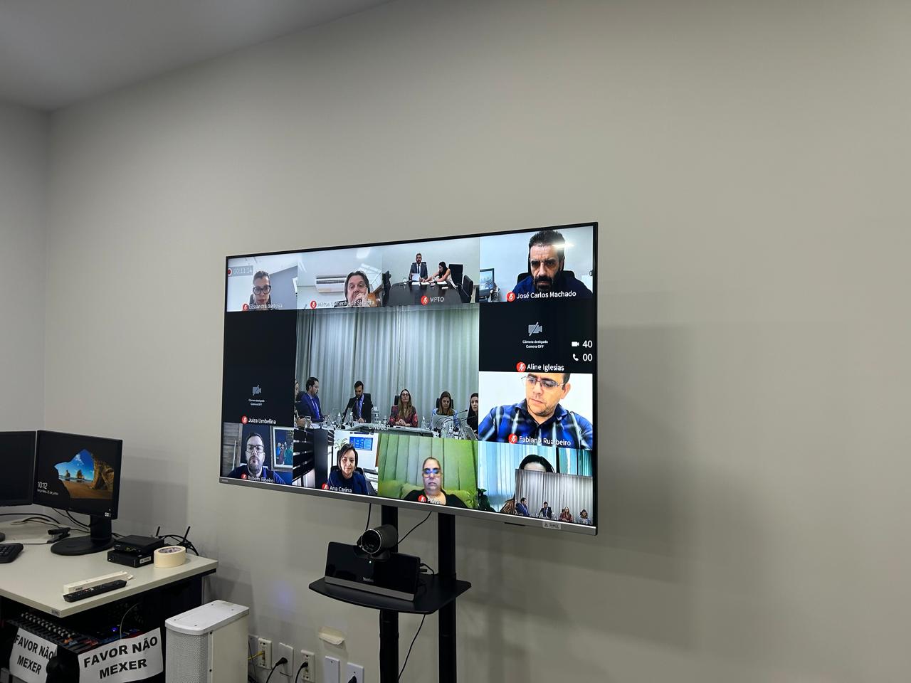 Fotografia colorida que mostra uma tela de televisão com vários quadradinhos onde há pessoas falando em reunião