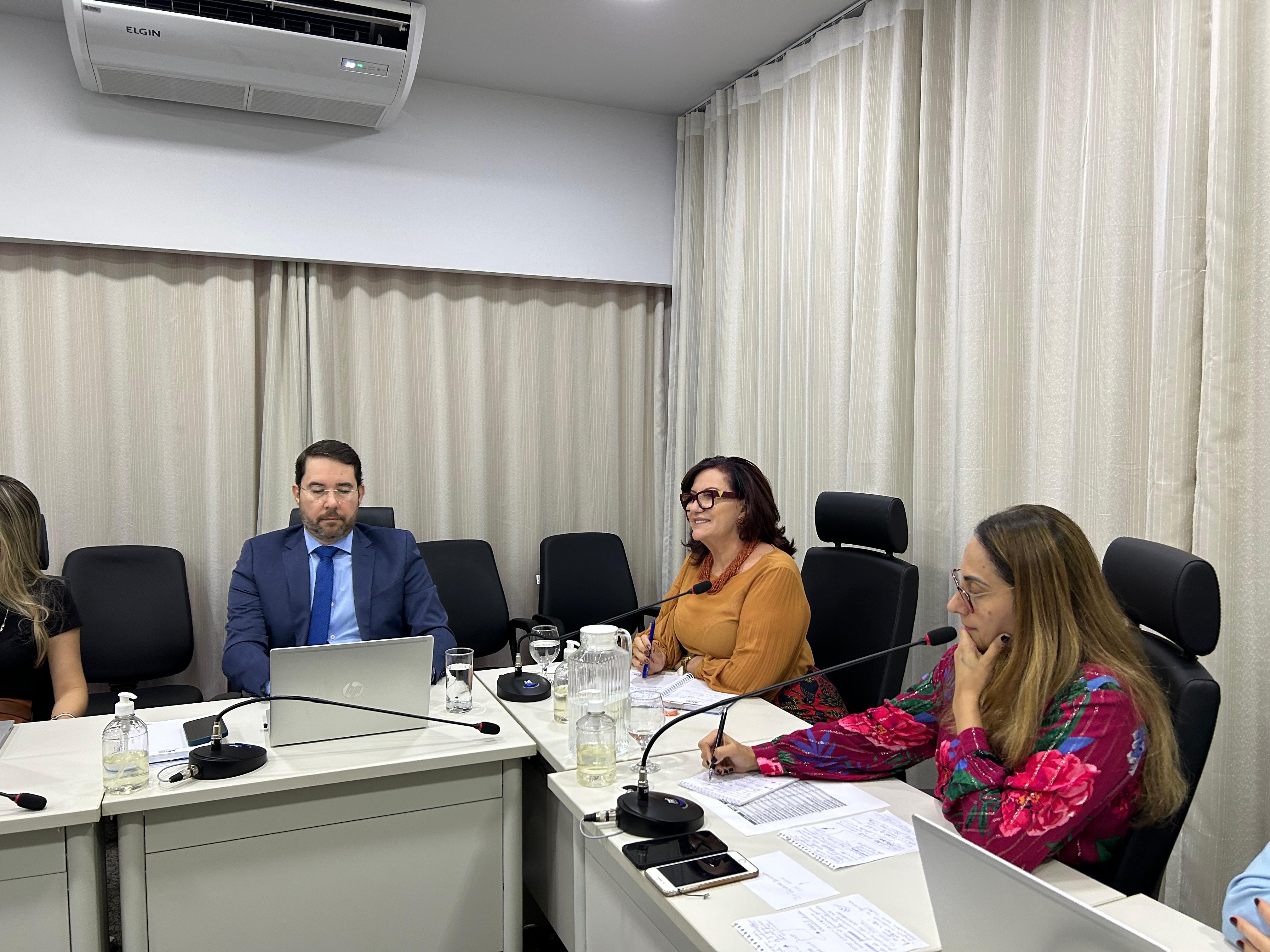Fotografia colorida que mostra uma mesa com três autoridades - sendo duas mulheres brancas e um homem branco de barba. Elas usam roupas coloridas, laranja e lilás com flores, ele usa terno e gravata azul. Os três estão em uma mesa de reuniões que tem microfones e copos à frente. 