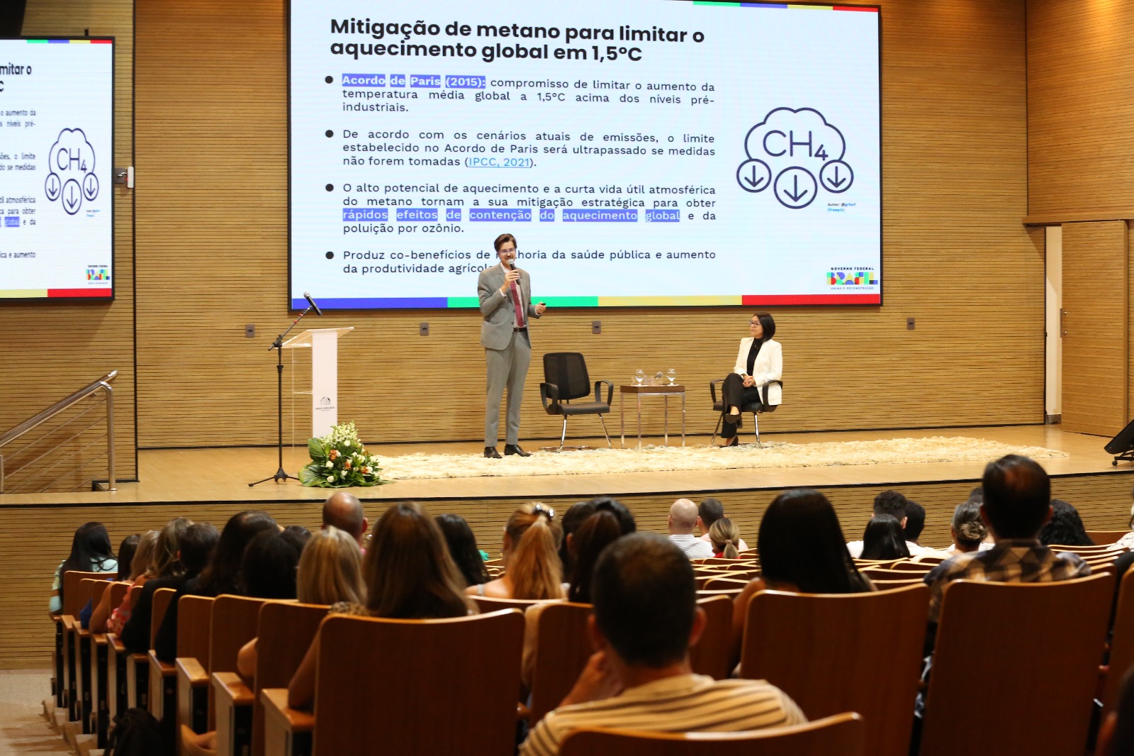 Imagem aberta do auditório, com detalhe de parte do público e, ao fundo, no palco, o palestrante em pé, falando ao microfone, e a mediadora sentada observando-o, e o telão com slide da palestra
