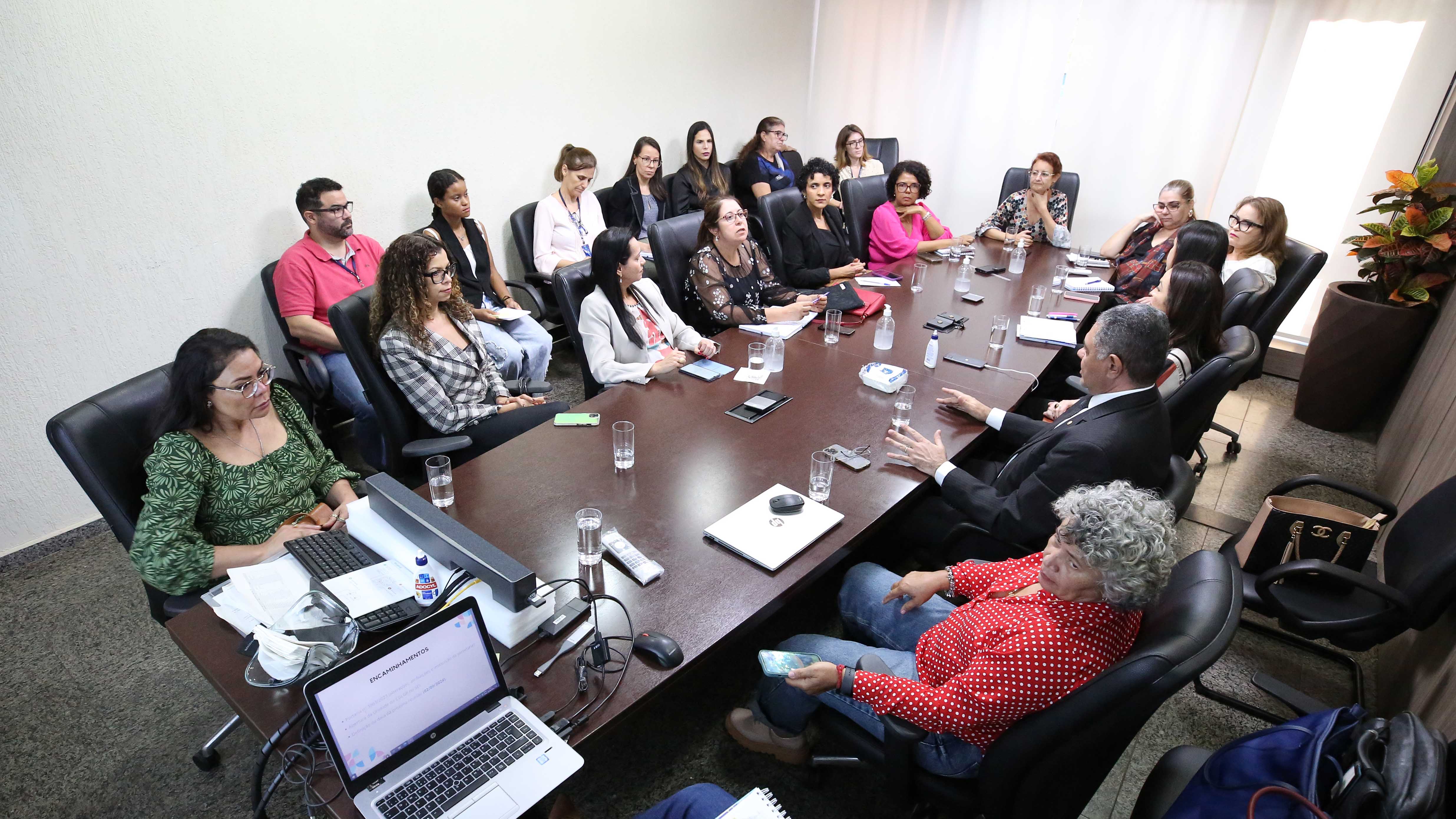 Fotografia colorida que mostra homens e mulheres sentados à mesa numa sala de reuniões