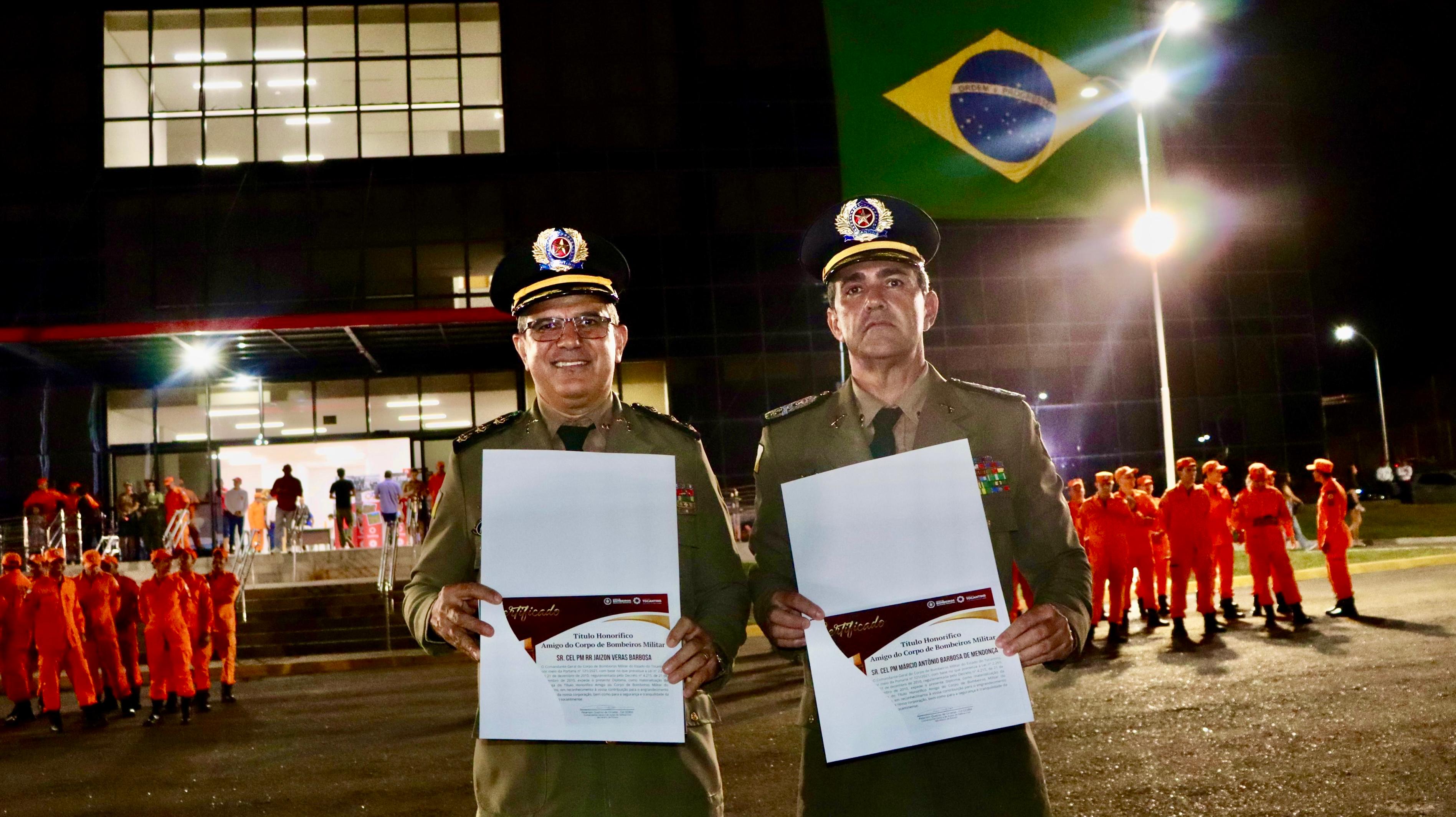 dois militares fardados posam para foto segurando diploma, ao fundo bombeiros militares com farda cor laranja estão em formação em frente ao prédio do quartel geral onde flamula a bandeira do brasil nas cores verde amarelo e azul