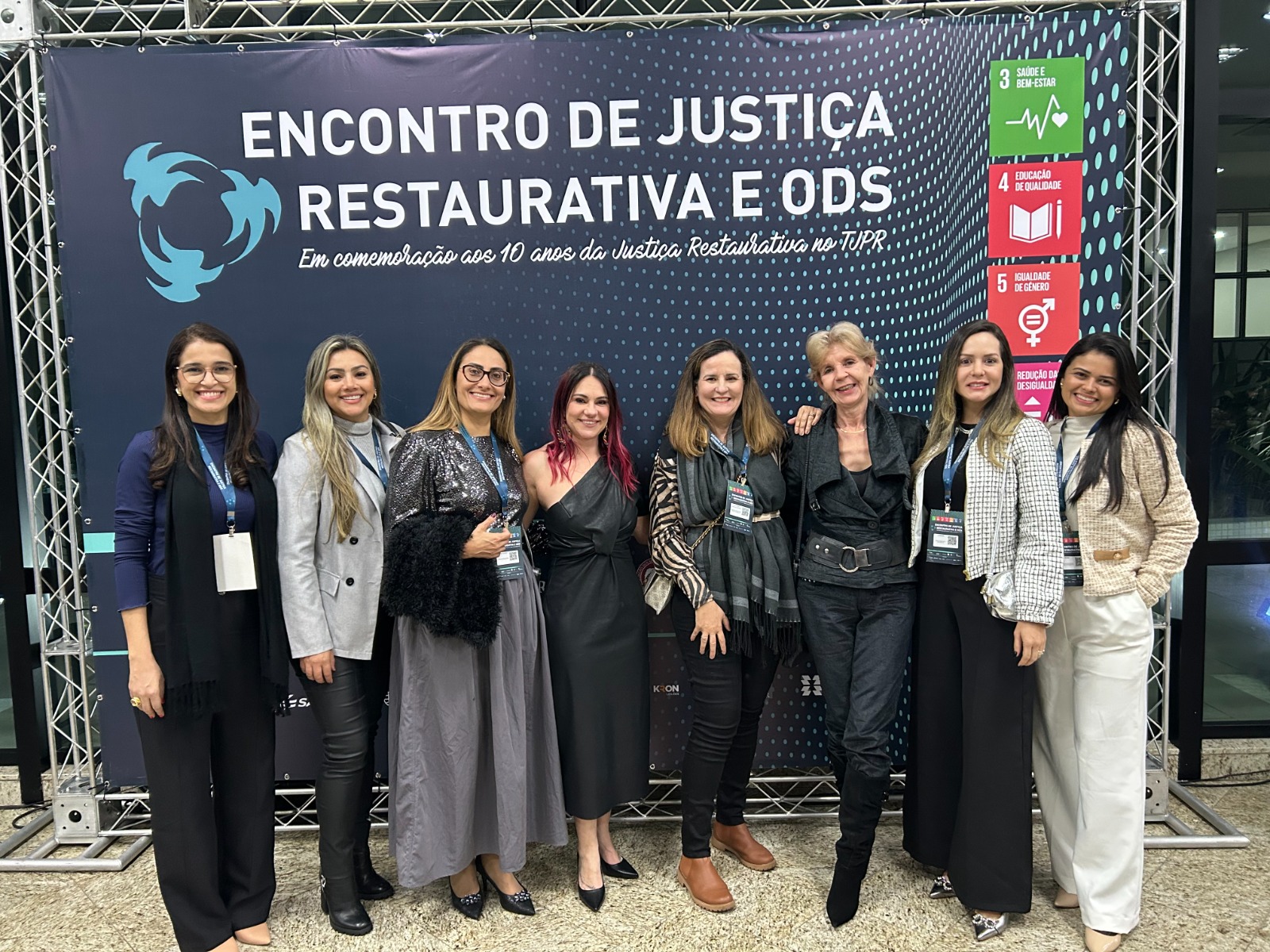 Oito mulheres estão em pé posando para foto e atrás tem um banner escuro