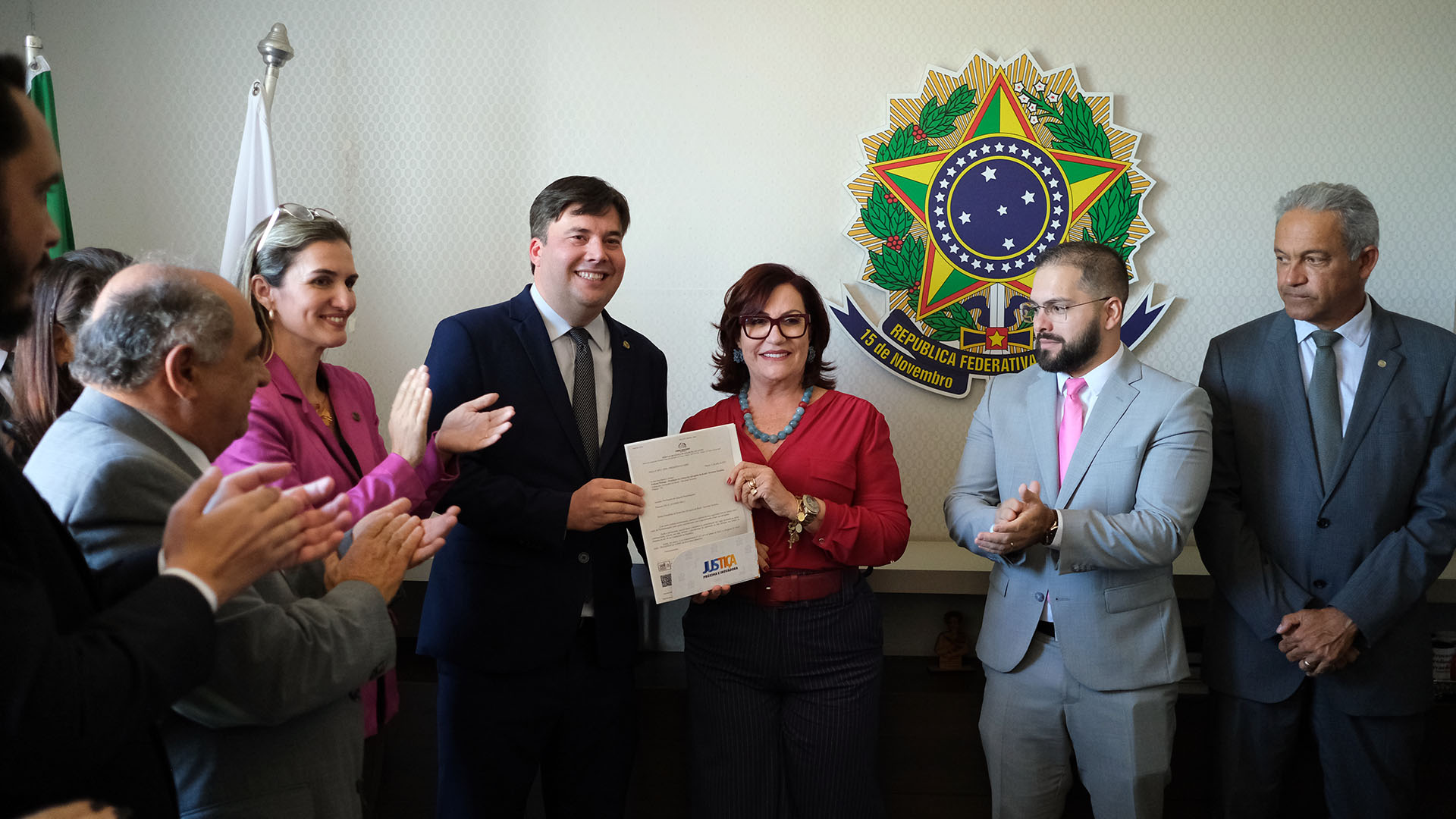 Pessoas estão em pé e algumas batem palmas enquanto outras duas seguram um documento