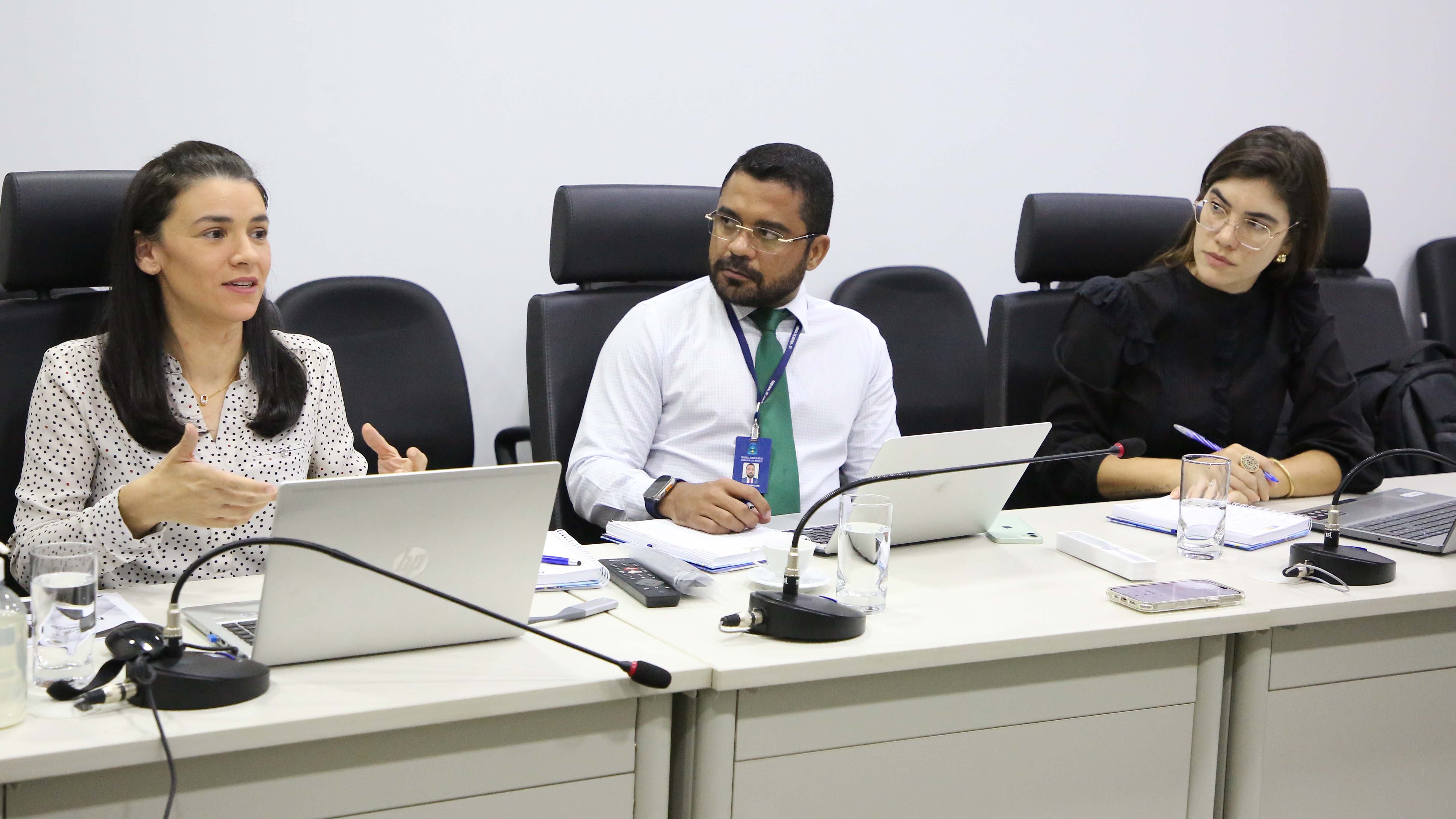 Mulher branca de cabelos pretos e lisos, na altura dos ombros, está sentada gesticulando enquanto fala. Ao lado dela, um homem moreno de camisa branca e gravata verde está de óculos a olhando. Na ponta, uma mulher branca, de camisa de manga comprida, também de óculos e cabelos lisos e longos a observa