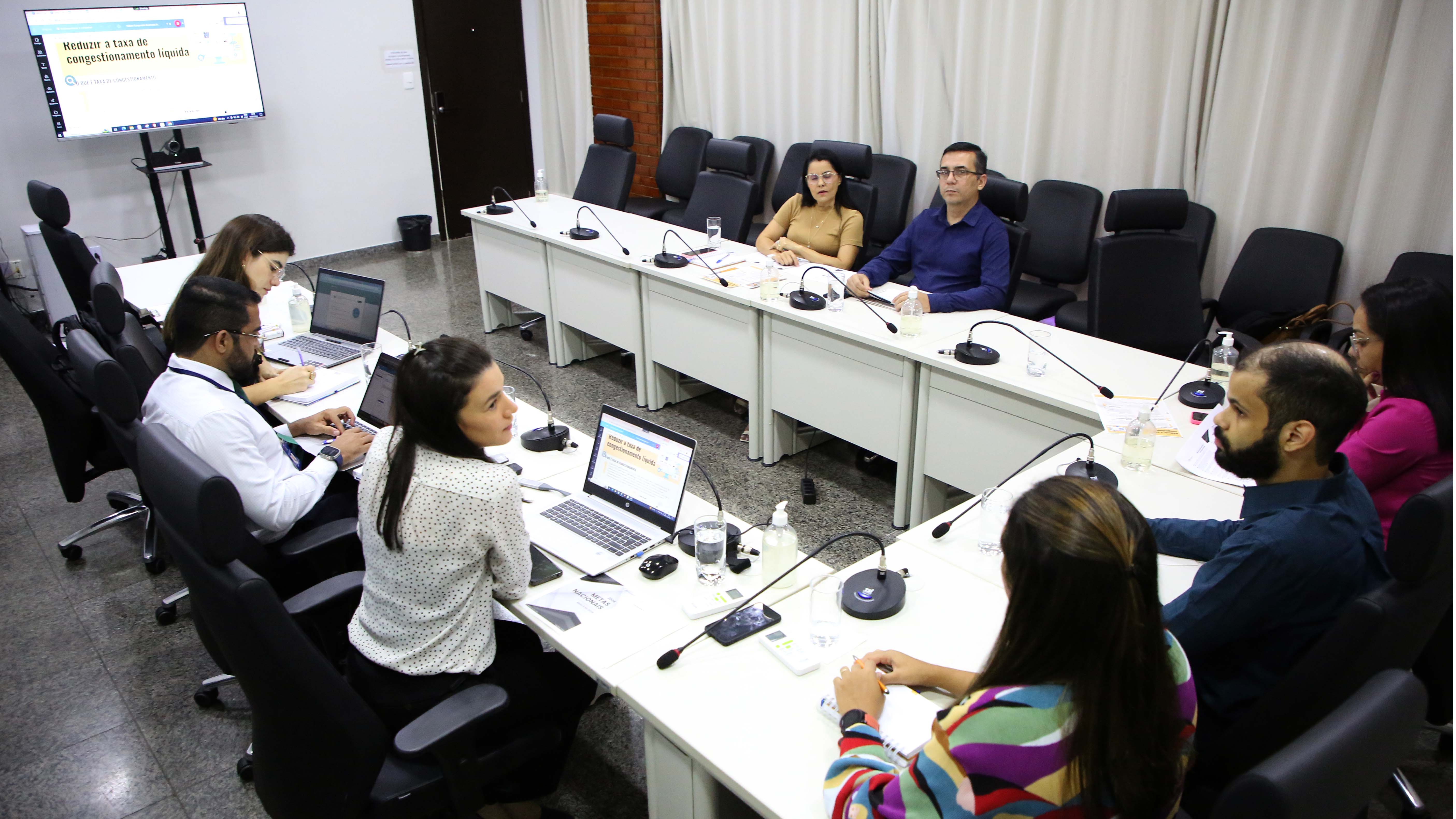 Homens e mulheres sentados em torno de mesa branca em formato U, com microfones individuais, durante reunião do TJTO, numa sala de paredes brancas com cortina branca. Ao fundo, uma tv projeta assunto da reunião, ao lado da tv tem uma porta de madeira