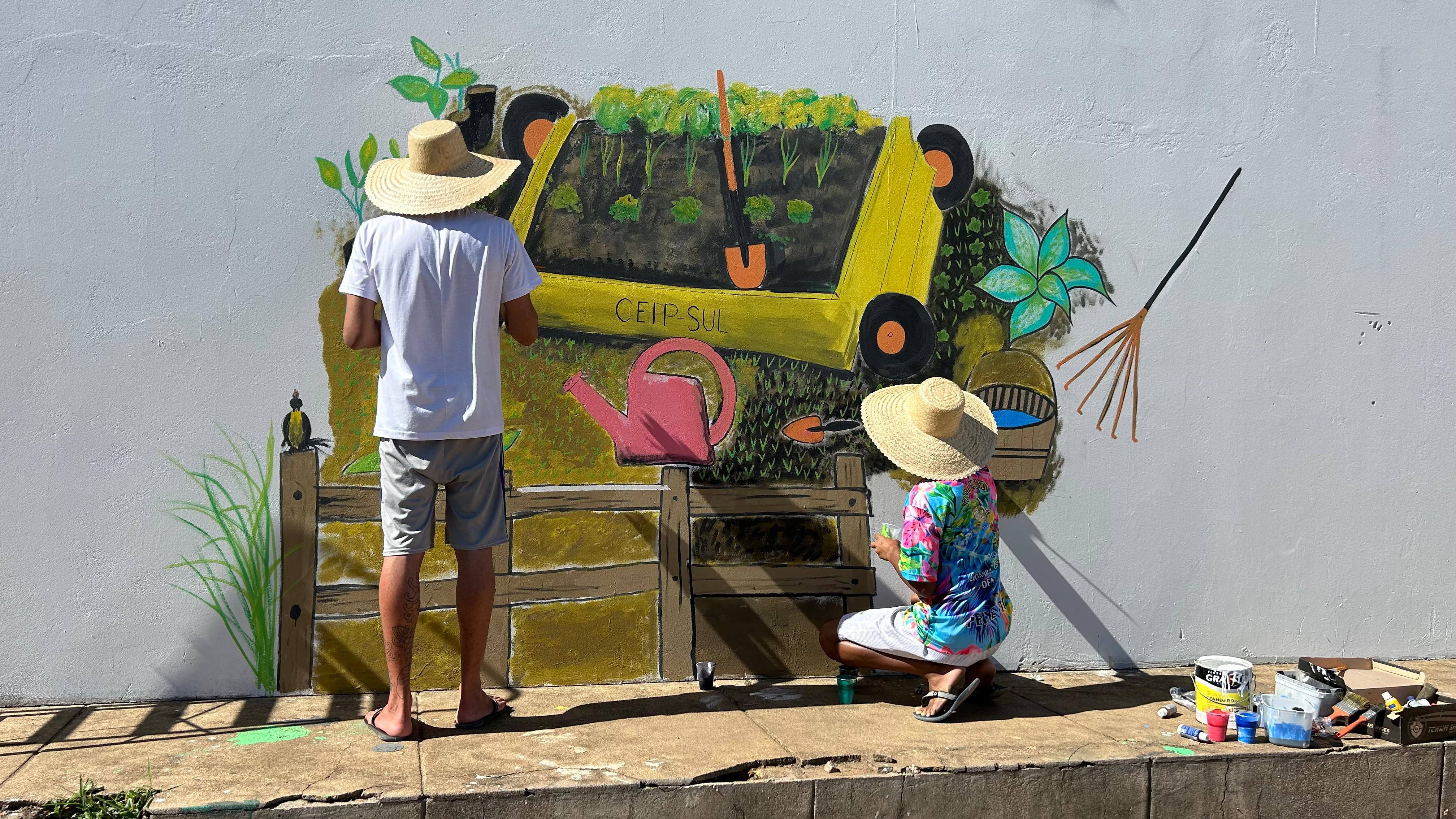 Imagem mostra painel desenhado na parede e duas pessoas com chapéu pintando