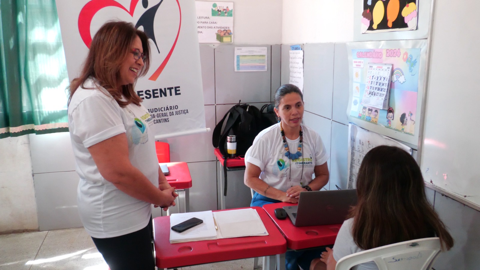 Imagem colorida que mostra uma mulher branca em pé e outra sentada atendendo uma criança de costas