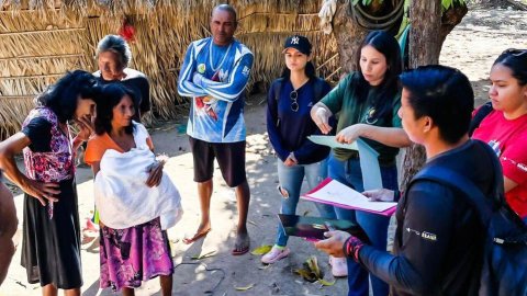 Imagem mostra uma mulher indígena segurando um bebê ao lado de outra que sorri; ao fundo pessoas da Funai