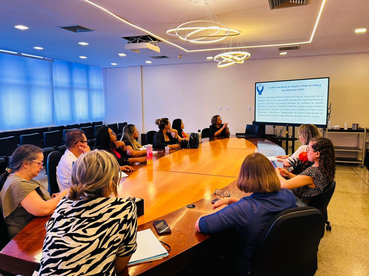 Uma sala com uma mesa grande em formato oval e 11 pessoas, sendo uma do sexo masculino, reunidas. Na imagem também aparece um telão com um texto projetado 