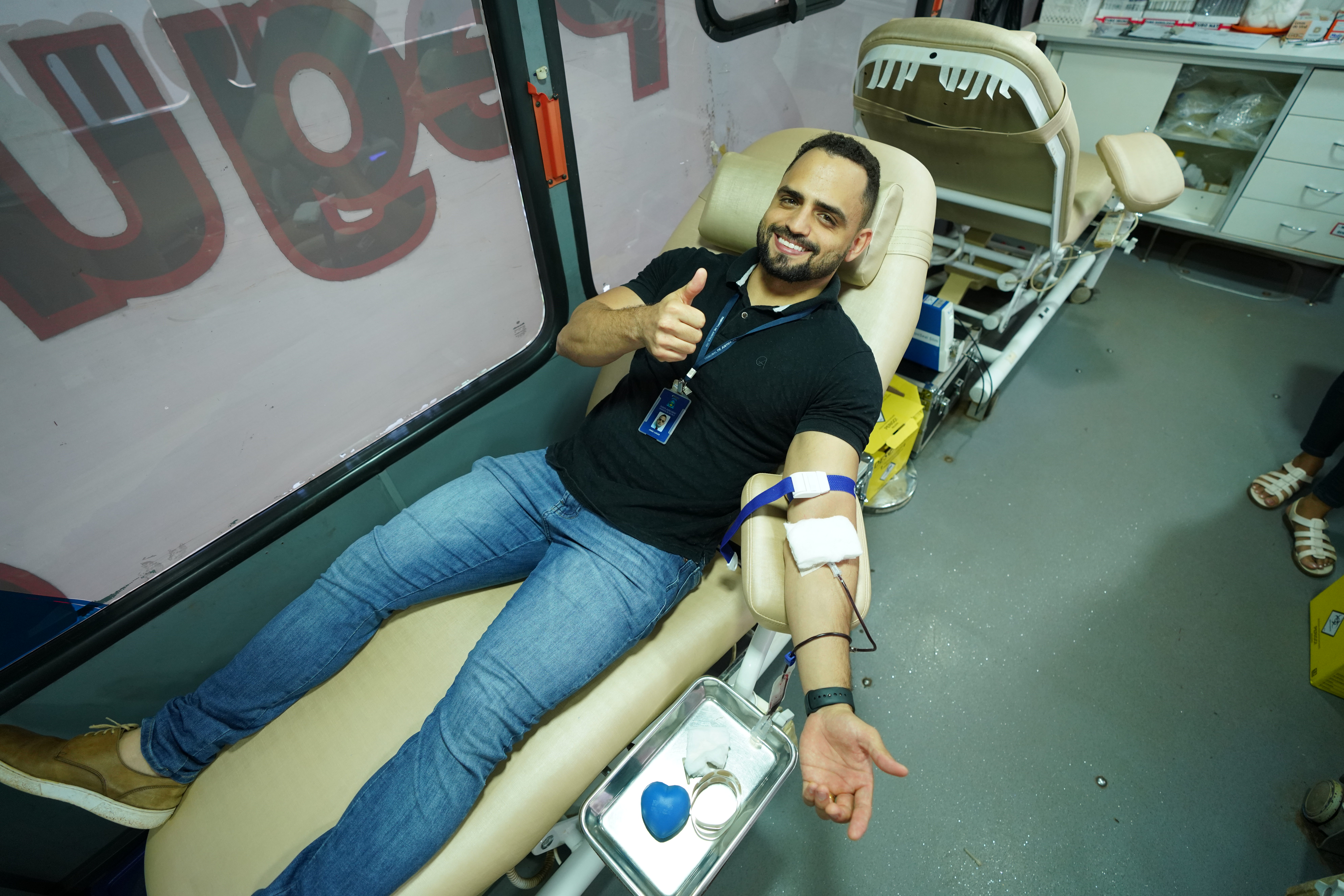 Fotografia colorida mostra homem deitado em cadeira de doação de sangue  e dando o sinal de joia 