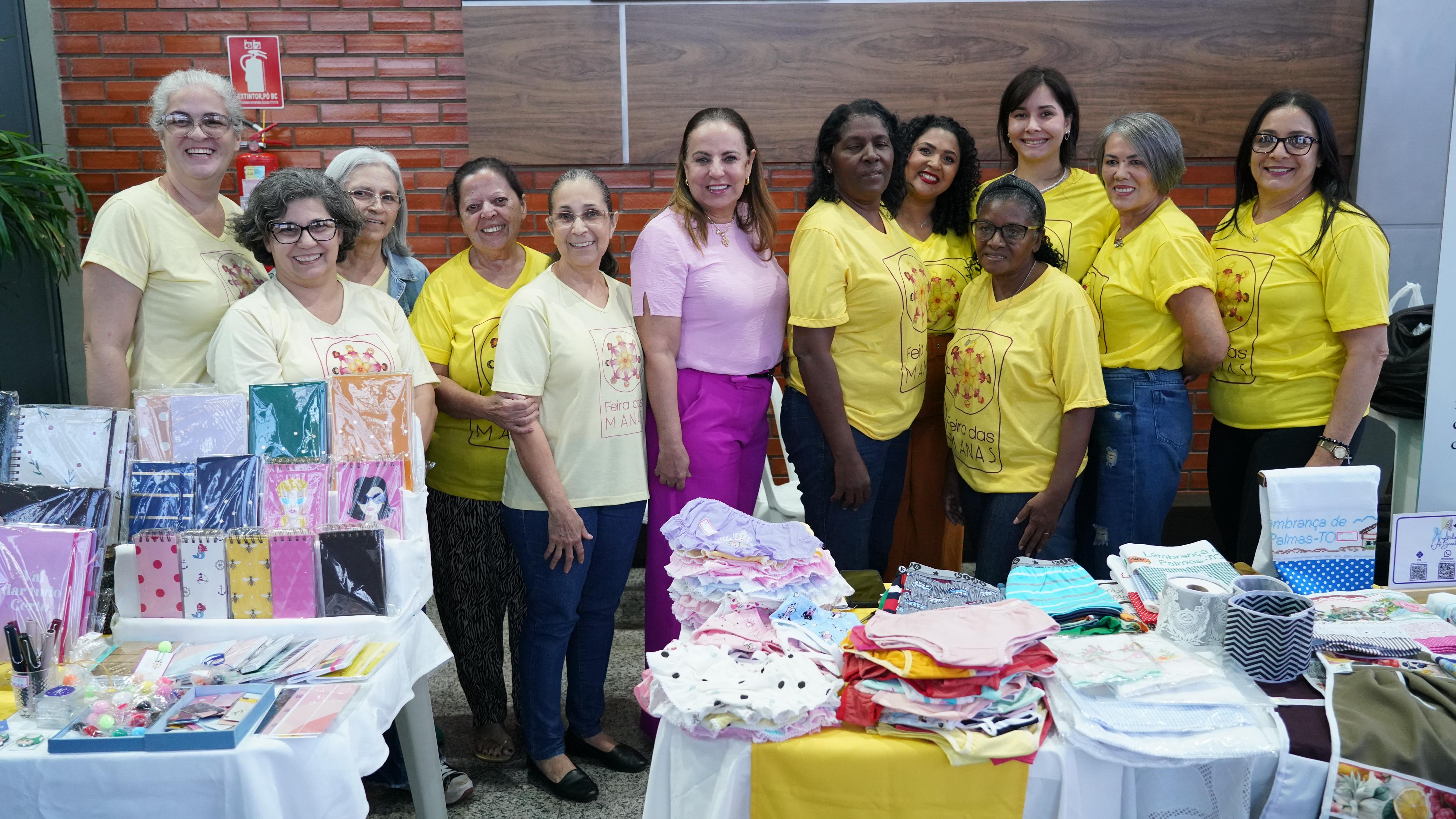 Mulheres em pé posam para foto atrás de mesa com produtos de artesanato