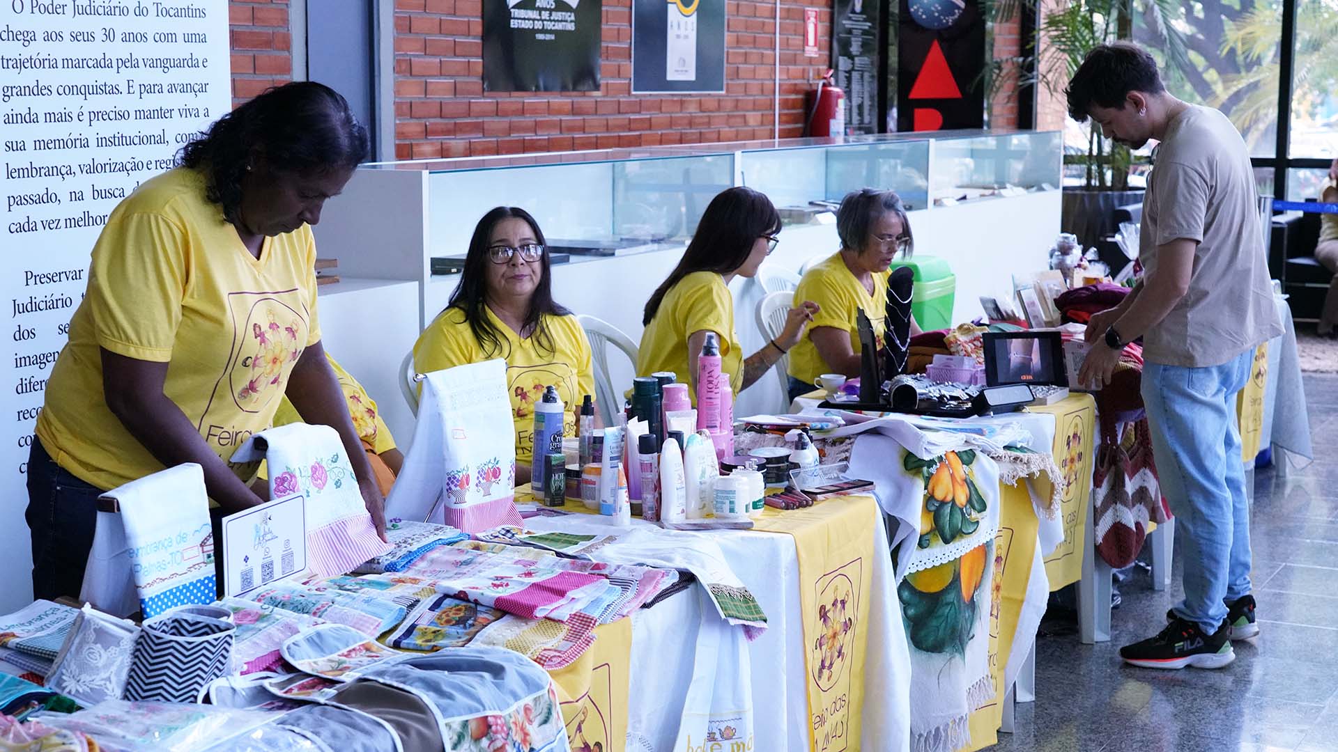 Mulheres de amarelo estão atrás de mesas onde expõem produtos de artesanato