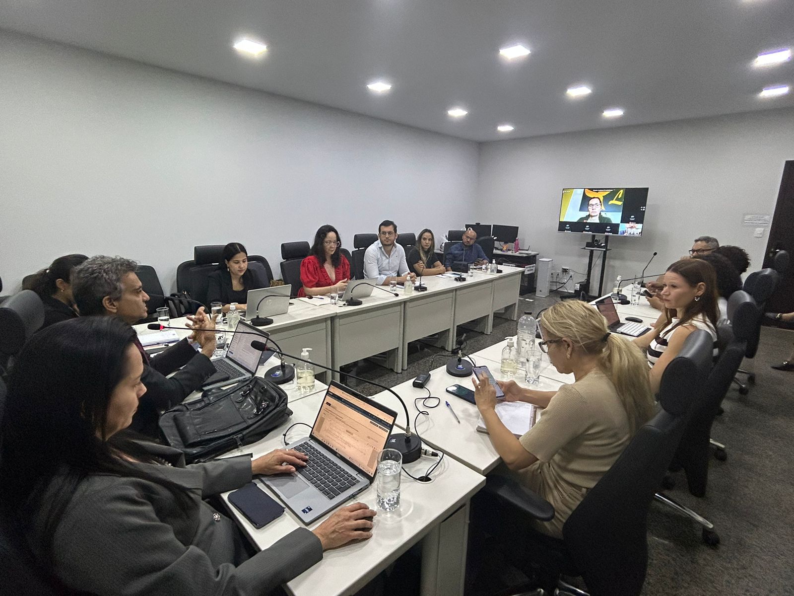 Homens e mulheres sentados em torno de uma mesa branca em formato de U durante reunião