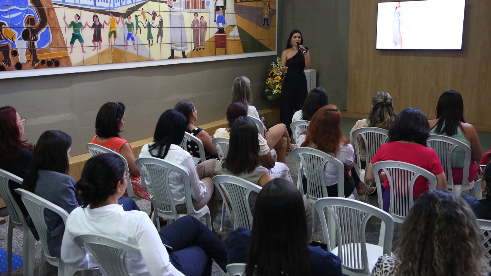Mulher em de preto em pé fala para uma plateia sentada em cadeiras de plástico branca; atrás uma televisão com uma apresentação