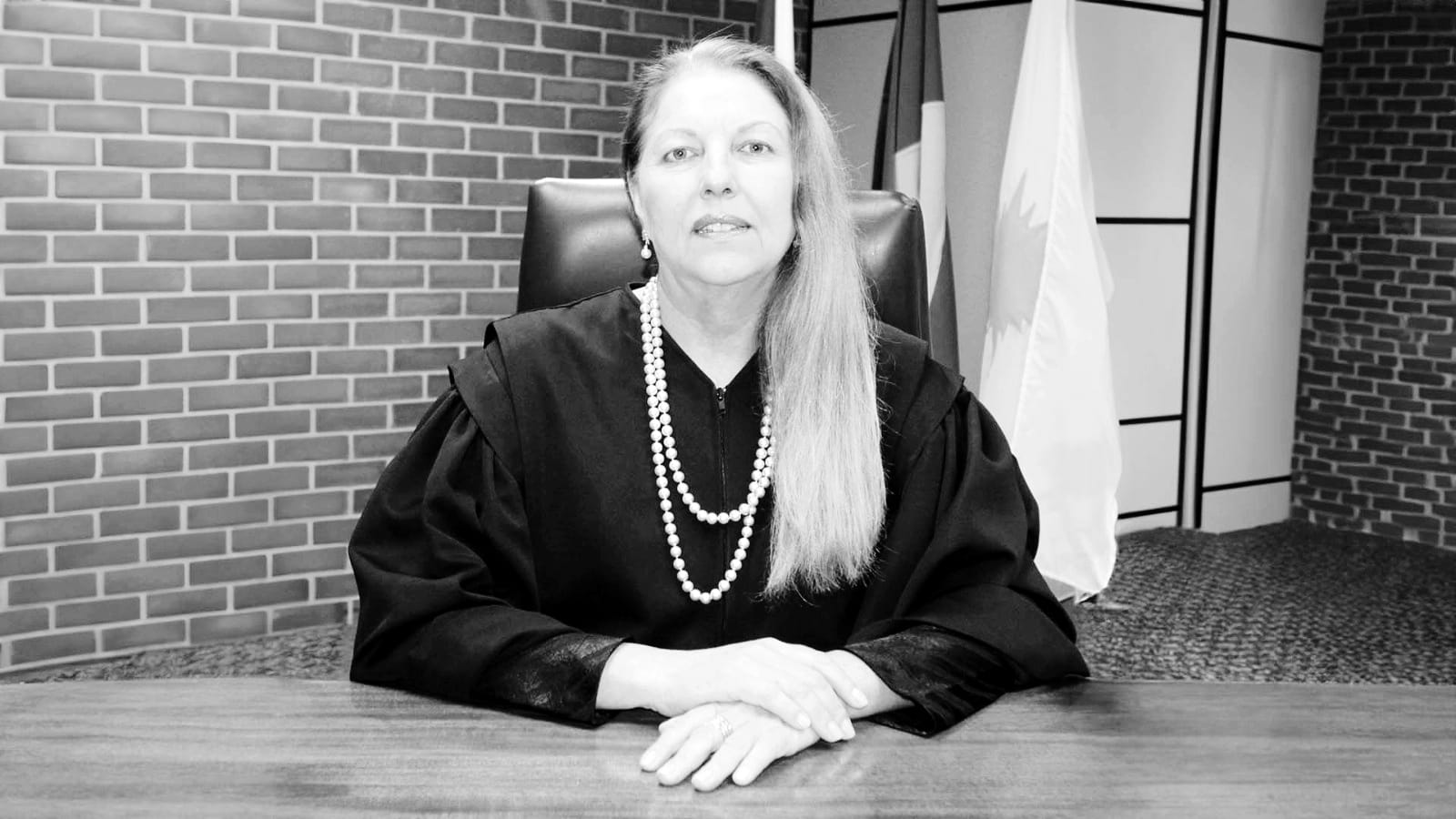 Foto em preto em branco de mulher branca de cabelos louros e longos, jogados no ombro. Ela é juíza, está de toga, vestimenta preta usada por magistrados, e está sentada em uma cadeira preta usando colar de pérolas. Ao fundo, parede de tijolinho exposto, com bandeiras na lateral direita