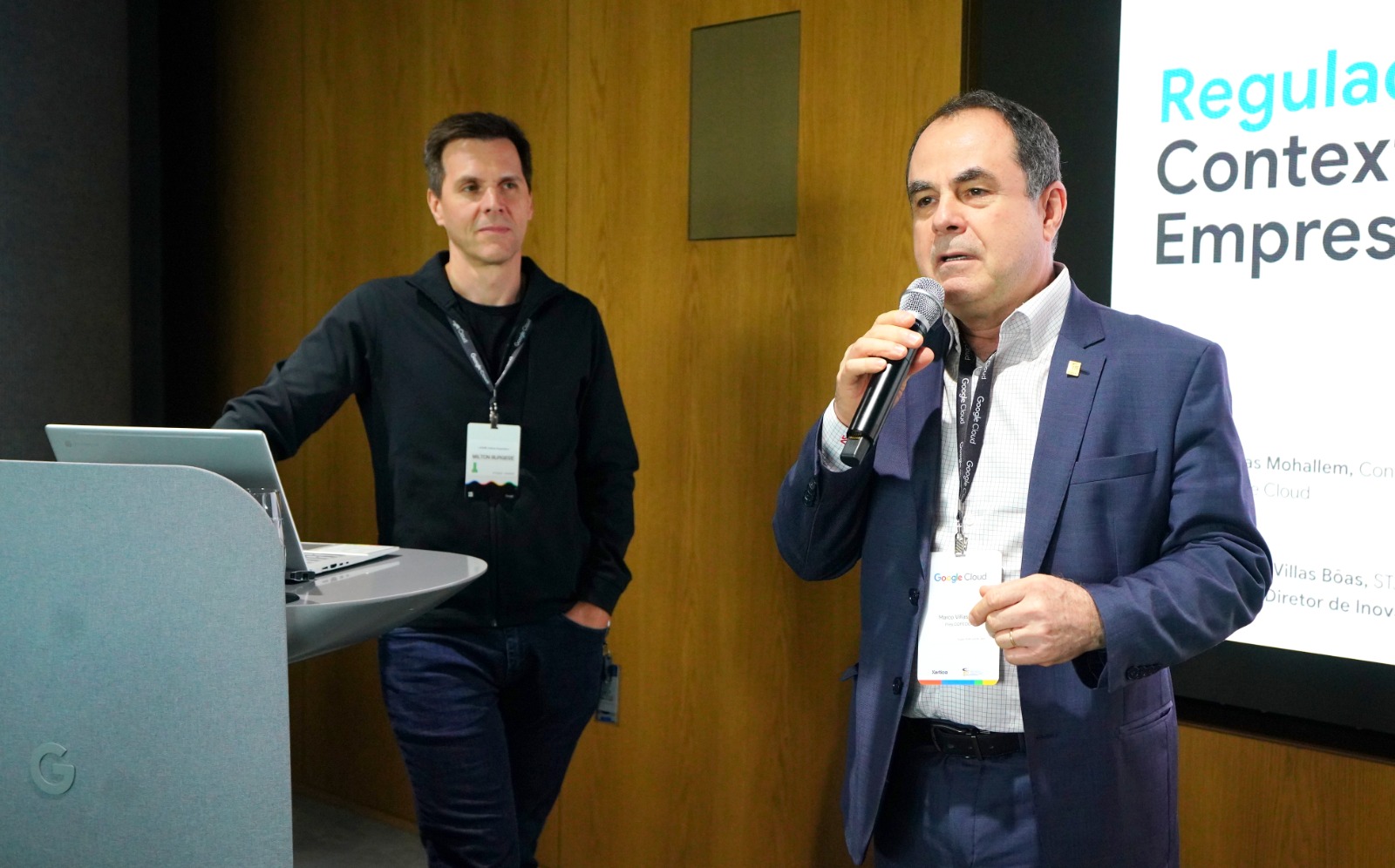 Dois homens brancos em pé; um está com microfone na mão e o outro está com a mão em uma bancada