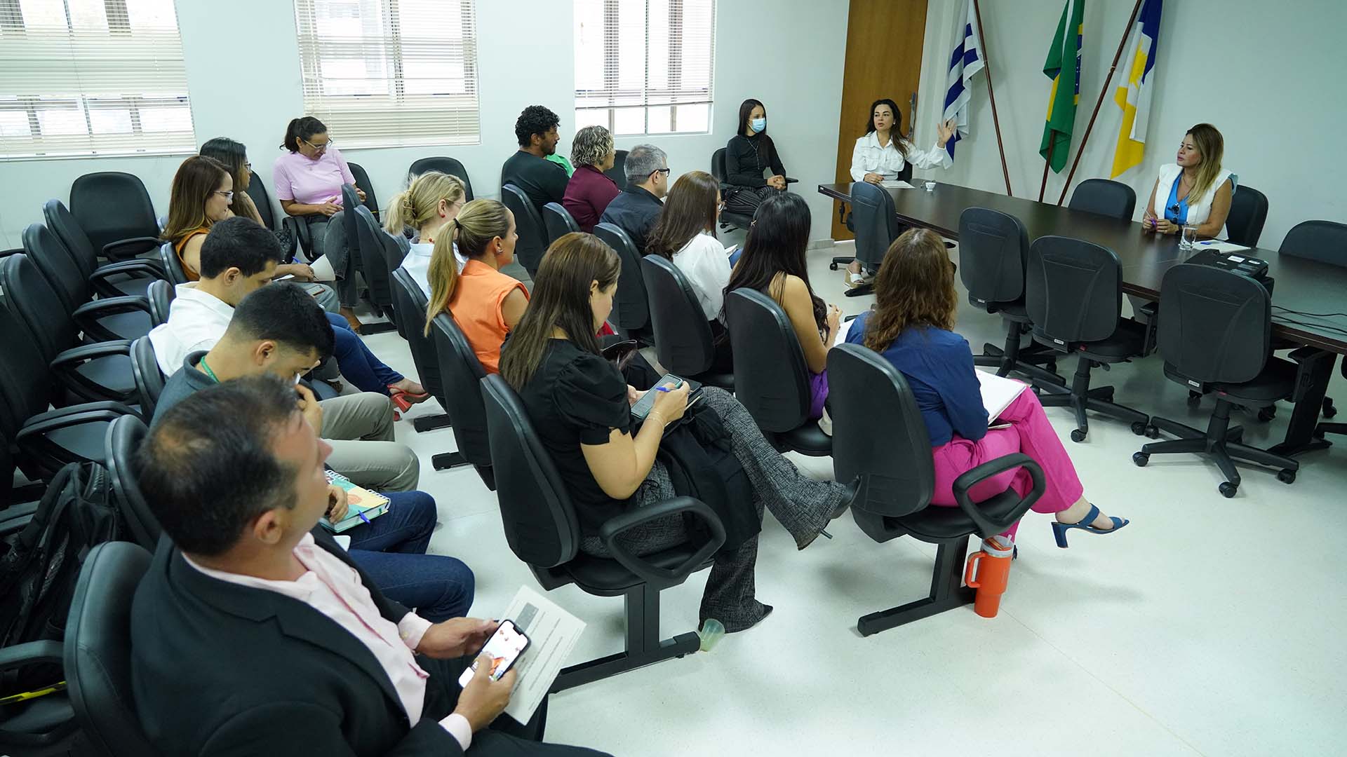Fotografia colorida que mostra homens e mulheres sentados observando uma mulher de branco que fala e, na mesa à frente, está uma mulher loira que observa