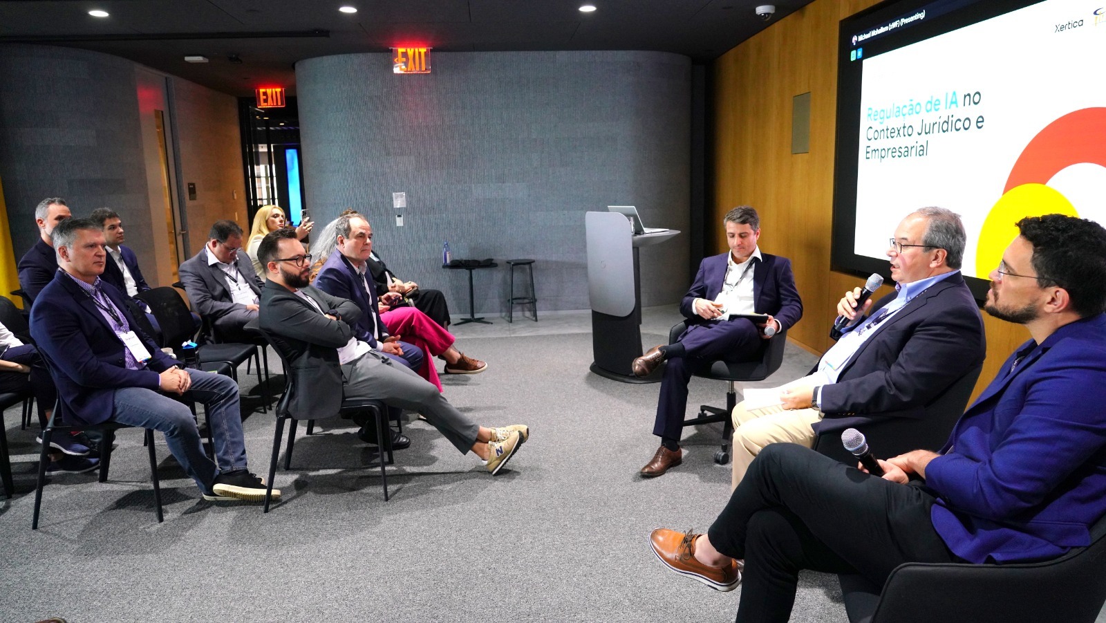Pessoas sentadas escutam palestra; atrás uma projeção colorida