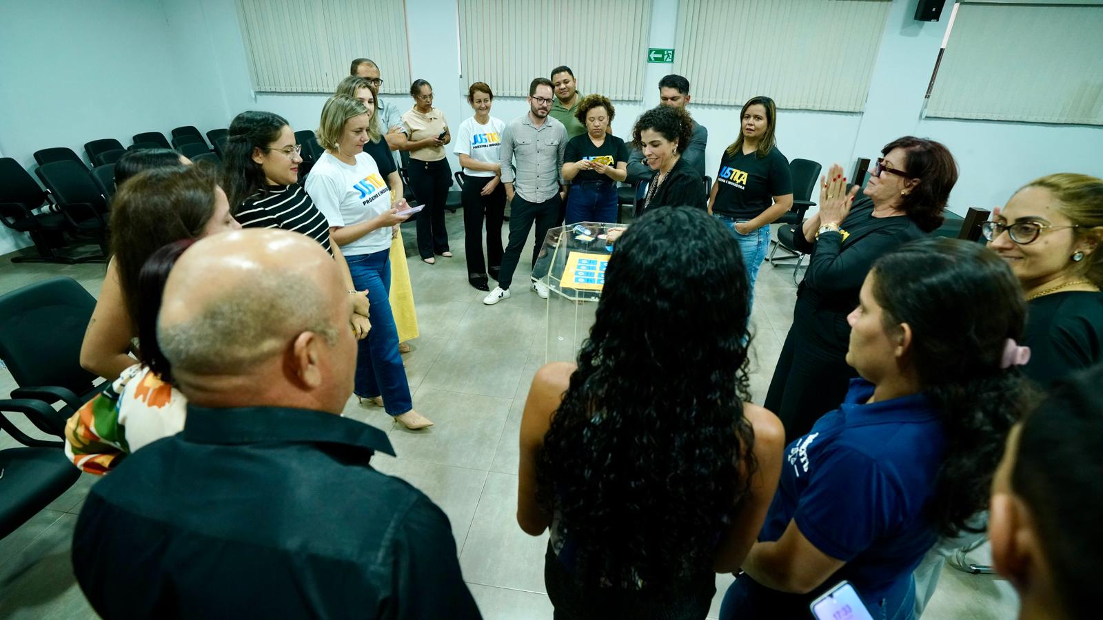 Várias pessoas em pé, com cartas na mão e uma mesa ao meio jogam o jogo da linguagem simplees