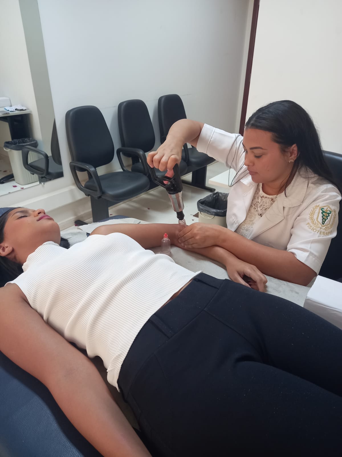 Fotografia colorida que mostra uma fisioterapeuta morena fazendo terapia manual numa jovem morena deitada. A jovem deitada em uma maca usa camiseta branca e calça preta