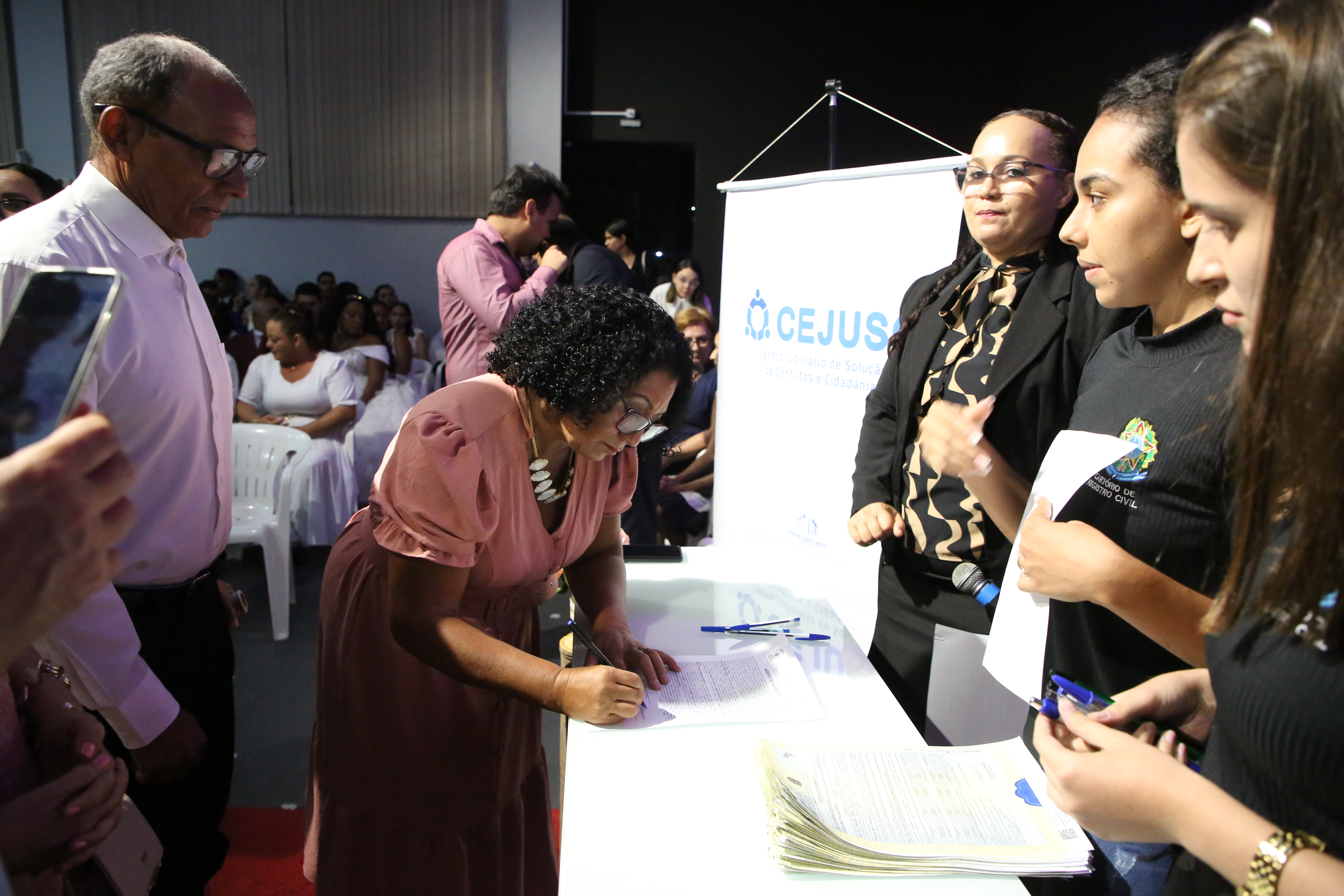 Ao lado do noivo, mulher assina Certidão de Casamento, diante de três mulheres trajadas de preto representado o sistema judiciário