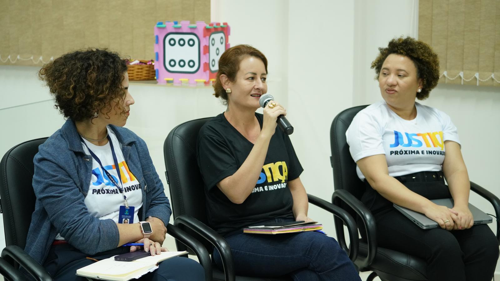 Imagem colorida que mostra uma mulher branca, usando camisa preta com a frase Justiça mais próxima e inovadora, falando ao microfone e sendo observada por duas mulheres