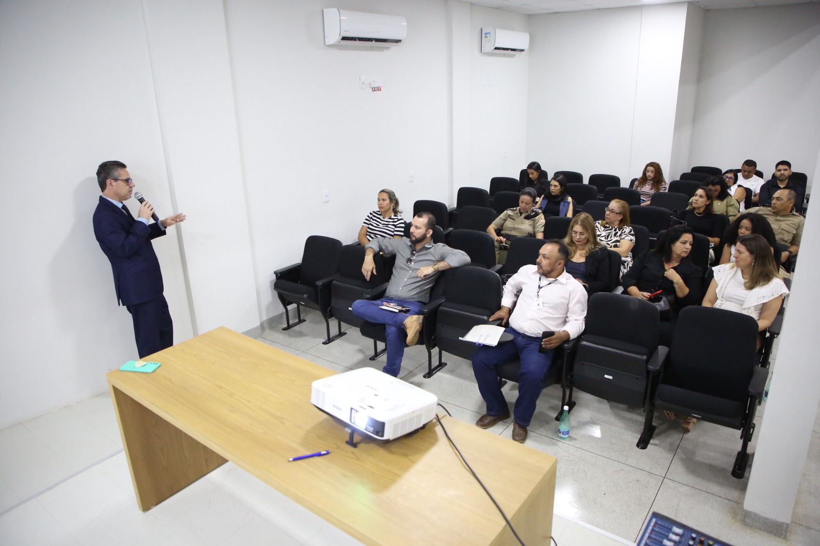 Homens e mulheres sentados em auditório enquanto homem pardo de terno preto encostado na parede branca fala ao microfone