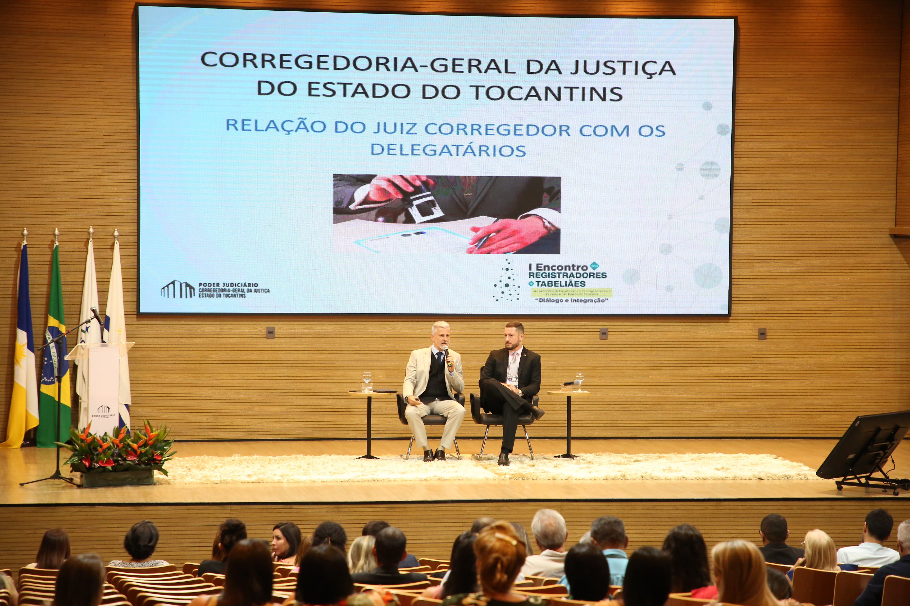 Homem branco, jovem, de cabelos brancos, vestido de terno claro, sentado ao lado de homem pardo, de terno azul escuro, fala ao microfone para plateia em auditório