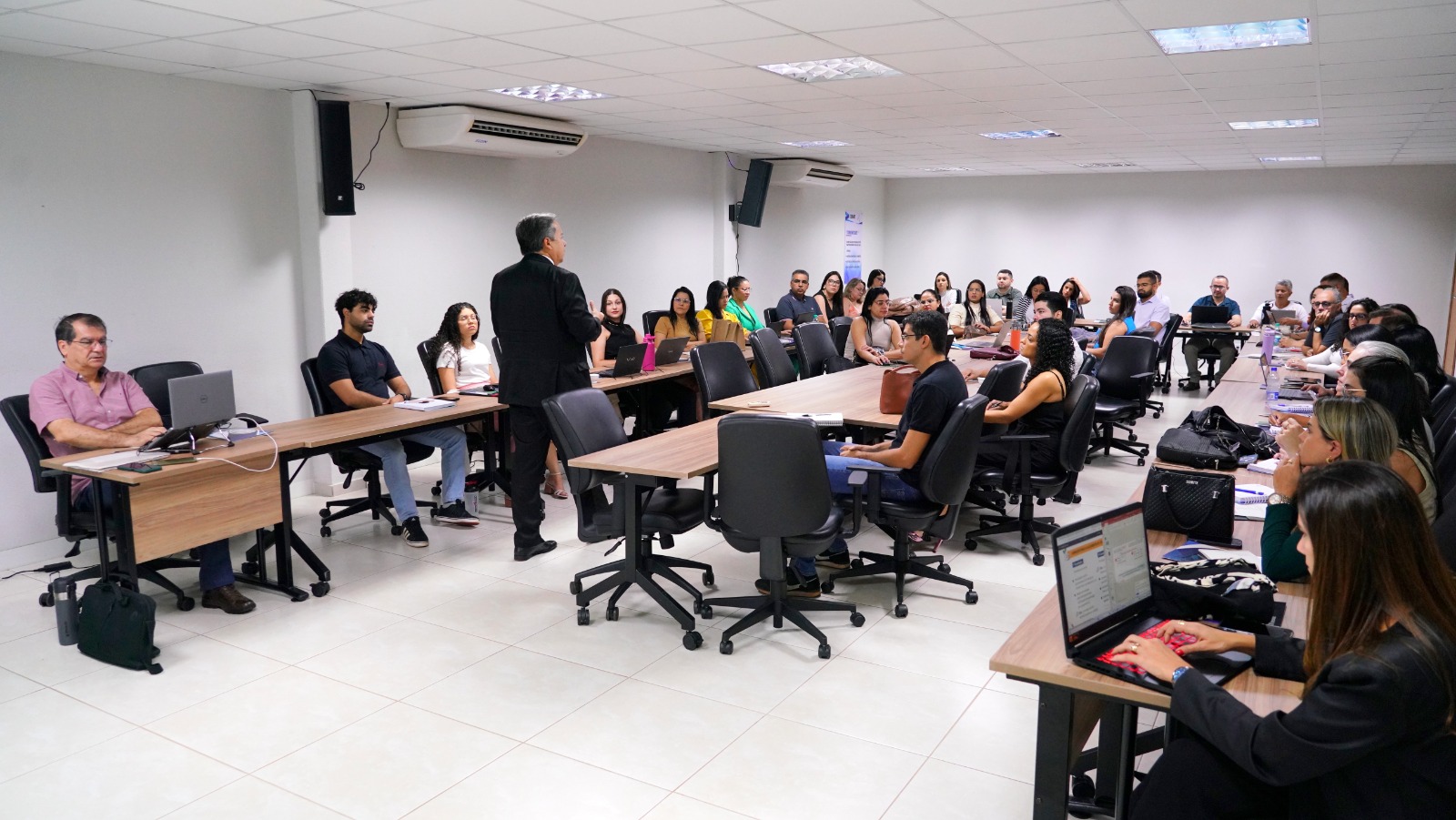 Sala de aula cheia de alunos com professor andando de costas, vestido de terno preto