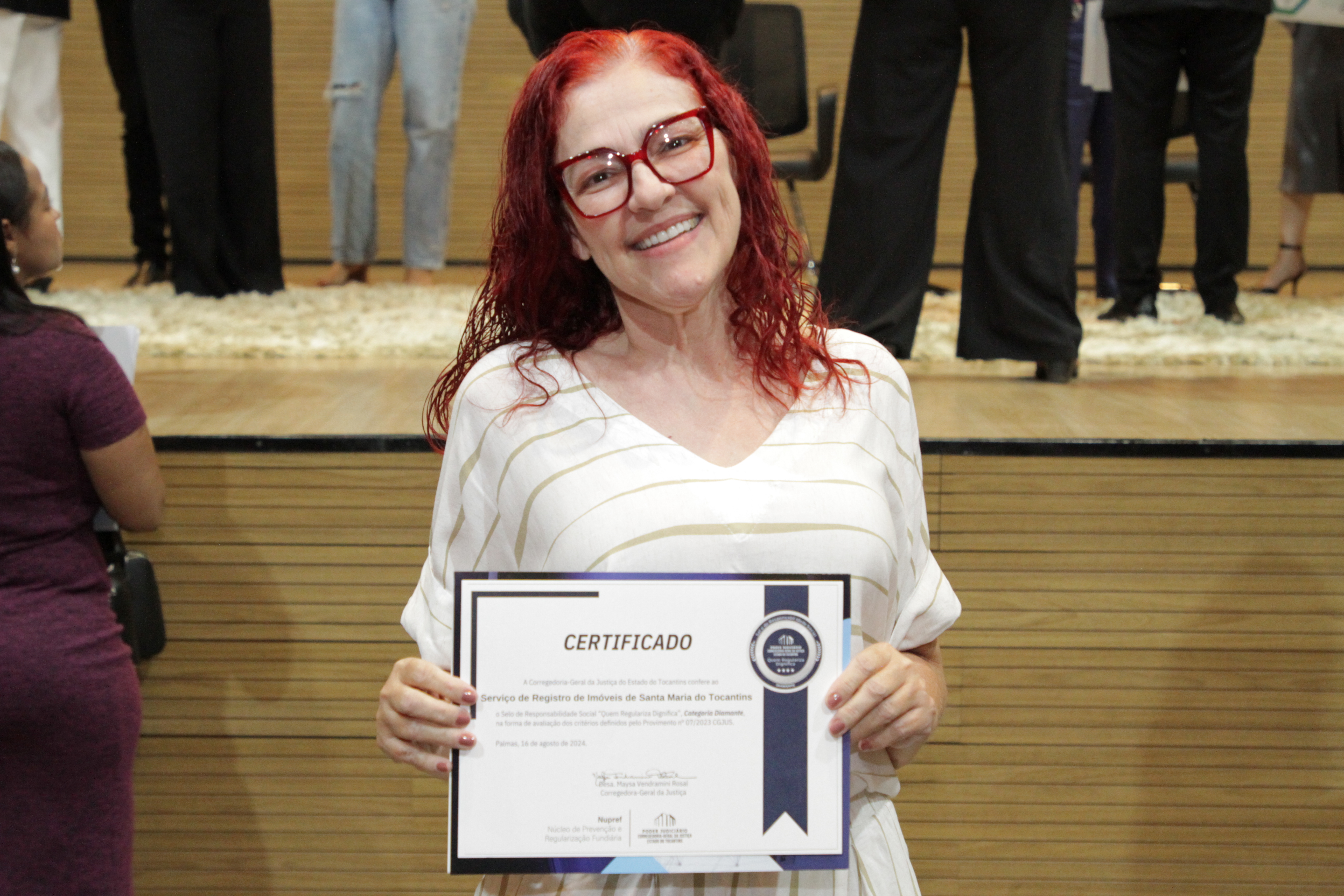 Fotografia colorida que mostra uma mulher branca, de óculos e camisa branca, segurando um certificado
