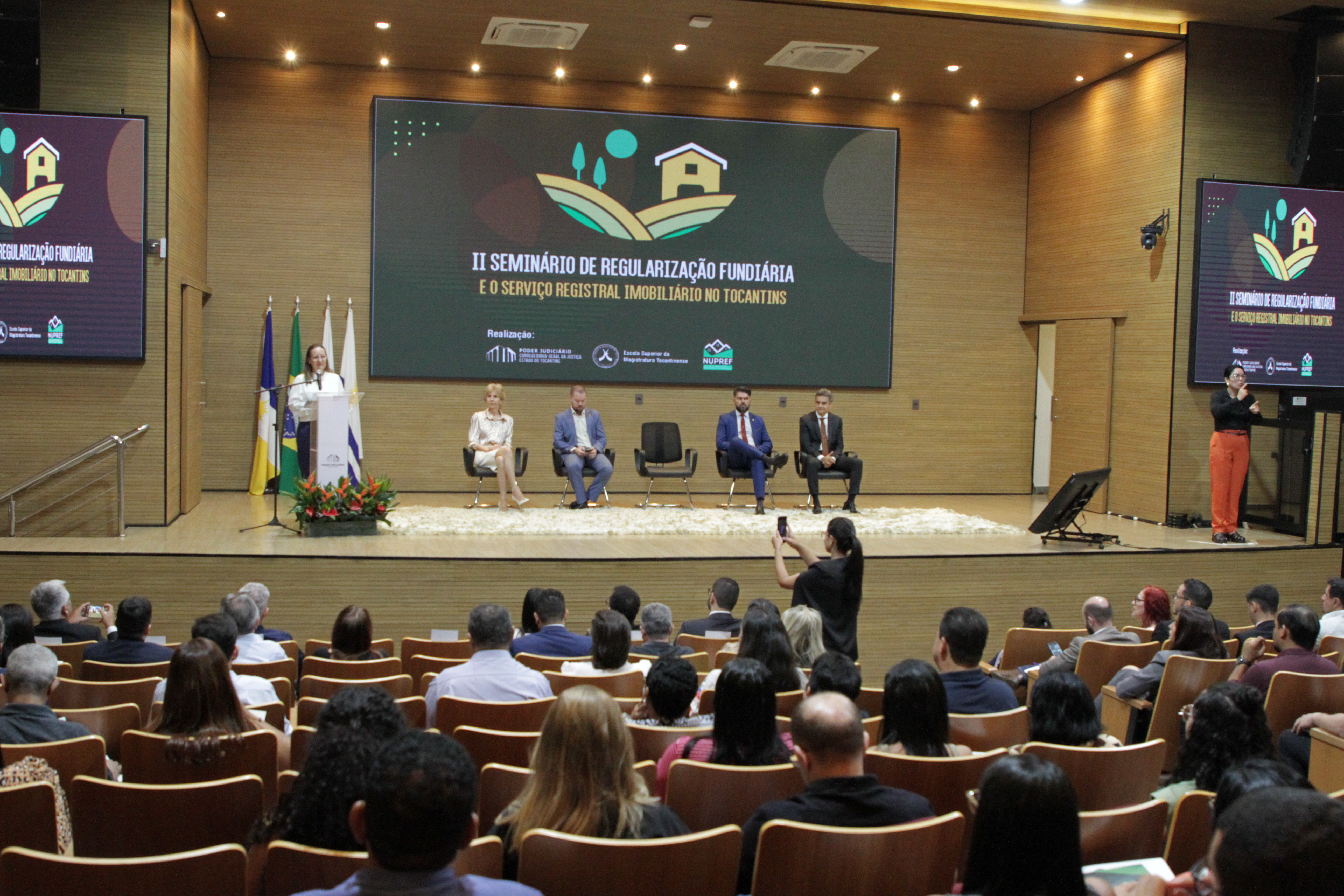 Fotografia colorida que mostra pessoas sentadas no palco num auditório e sendo observadas por uma plateia