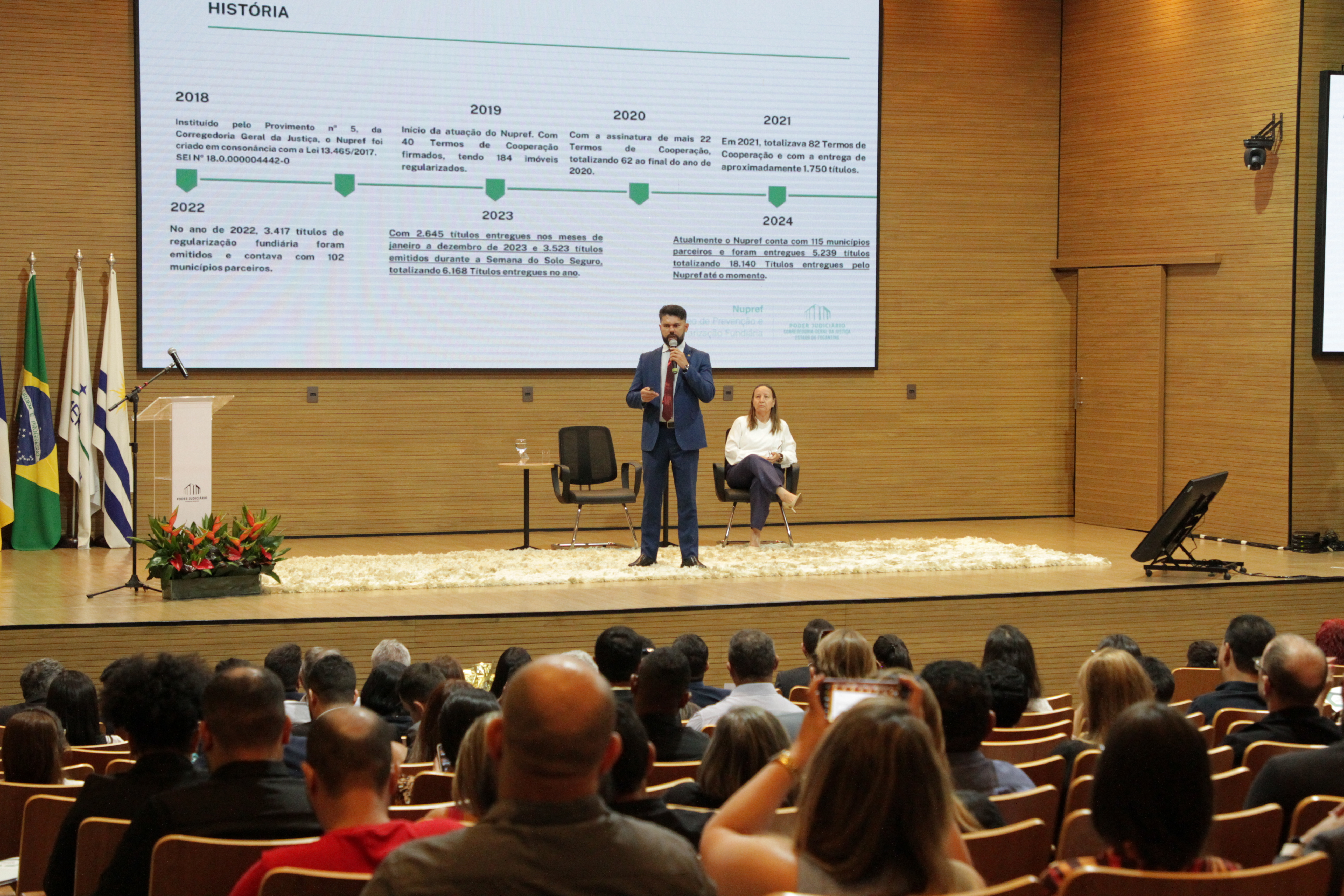 homem de terno escuro discursa ao microfone para plateia no auditório do tjto, mulher de blusa branca sentada ao palco também assiste