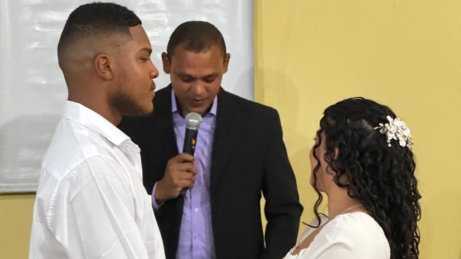 Homem negro, alto, casando com mulher parda, baixa, de cabelos longos cacheados, ambos trajados de branco. Juiz de paz celebra casamento, um homem negro, de paletó preto, camisa lilás, de microfone na mão