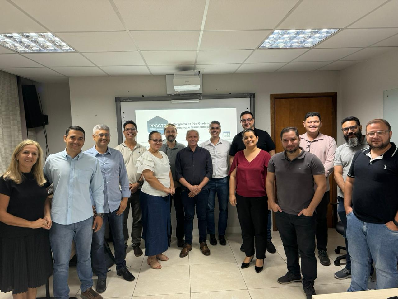 Homens e mulheres em pé em sala de aula de paredes brancas com telão ao fundo