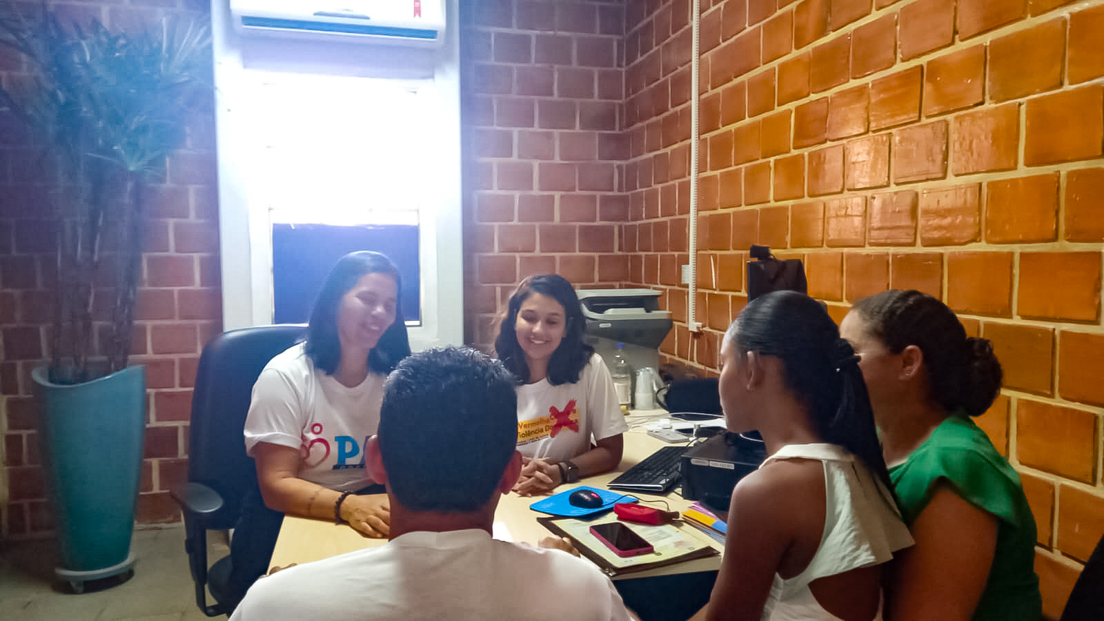 Duas mulheres atendem cidadãos(ãs) em sala de tijolinho exposto. Elas estão sentadas de frente, vestindo camisa branca, enquanto o casal e a filha estão de costas, ele e a filha de camisa branca e a mulher de camisa verde