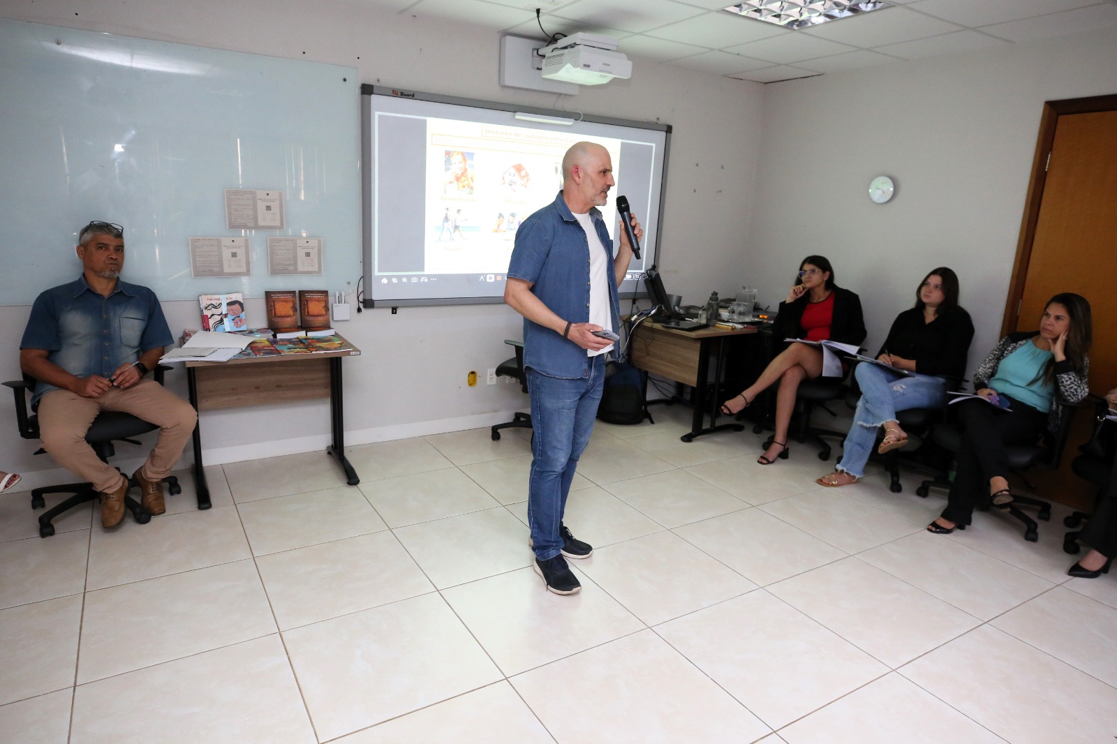 Em uma sala de aula, um homem branco, de cabelos e barba brancos, trajando calça e camisa jeans e camiseta branca, está em pé, de frente, falando ao microfone, à sua direita três servidoras estão sentadas lhe observando, à sua esquerda um servidor e ao fundo um quadro com uma imagem projetada.