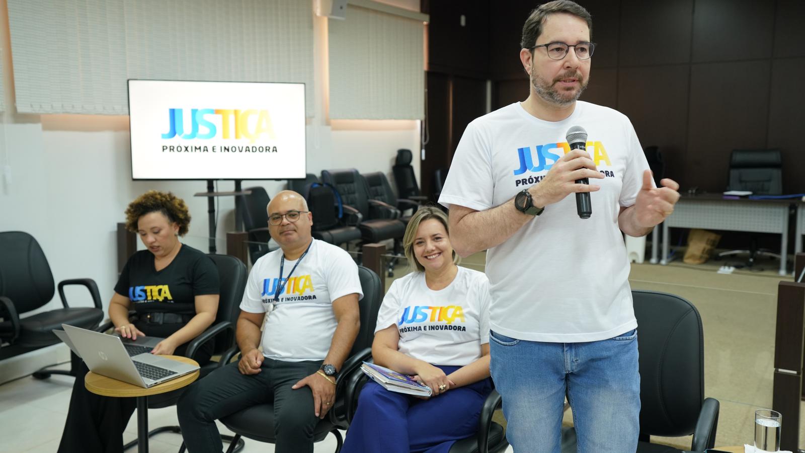 Fotografia colorida que mostra um homem branco discursando e duas mulheres observando. Todos usam camiseta do Justiça Mais Próxima 