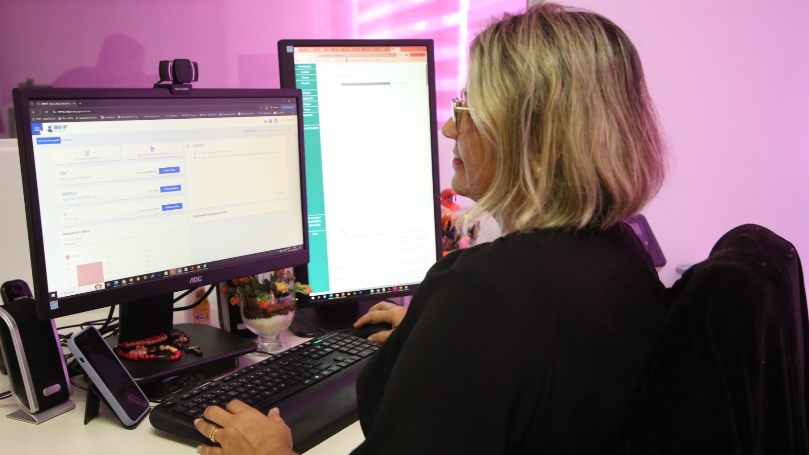 Mulher branca de cabelos lisos, louros, na altura dos ombros, opera computador em duas telas. Ao fundo parede branca com persiana horizontal, com reflexo de luz rosa na parede