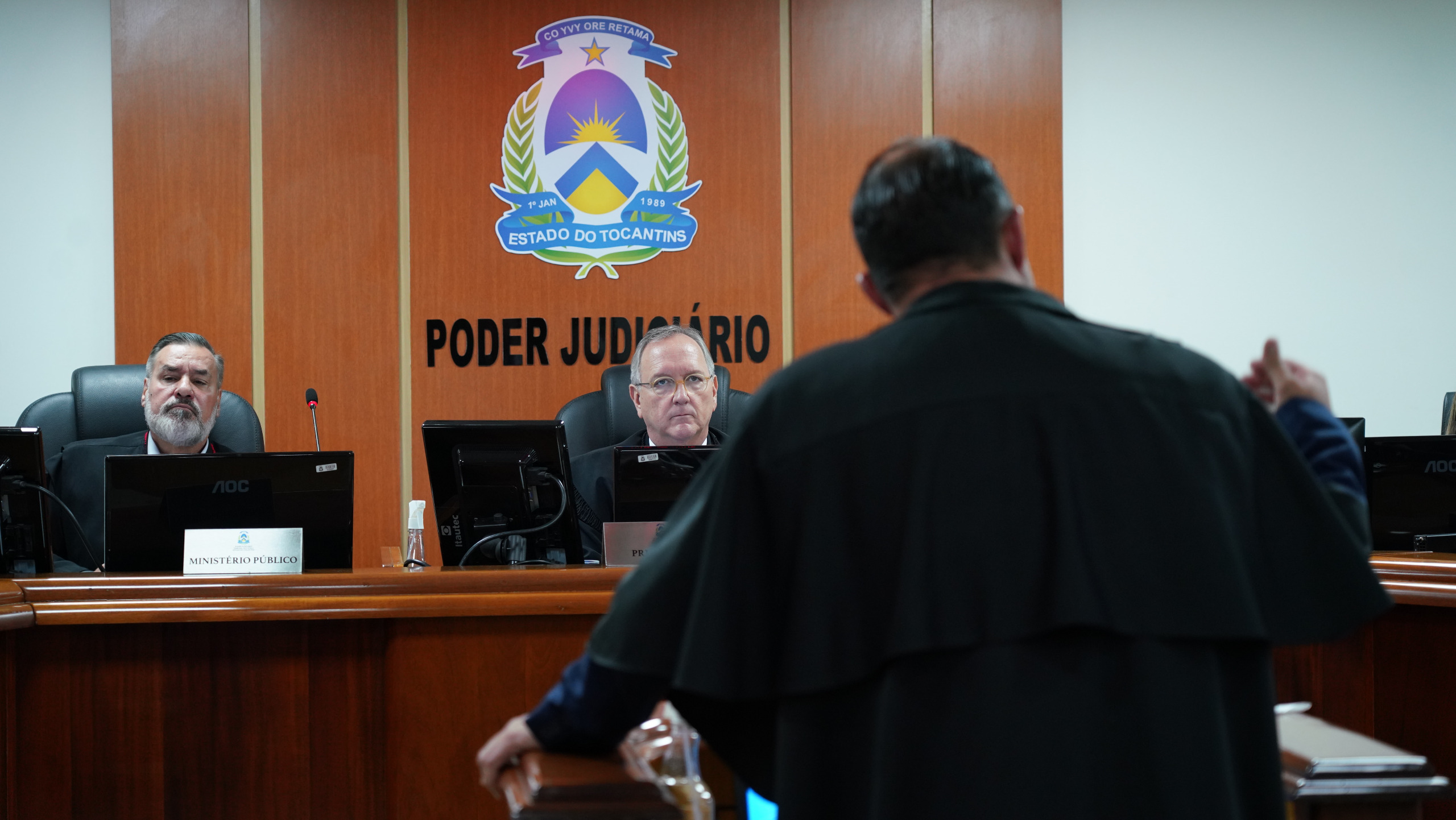 Homem de costas trajando toga preta de advogado no pleno do TJTO. Ao fundo, desembargadores sentados à mesa de madeira, tem um brasão do Poder Judiciário no centro da parede com acabamento em tábuas de madeira