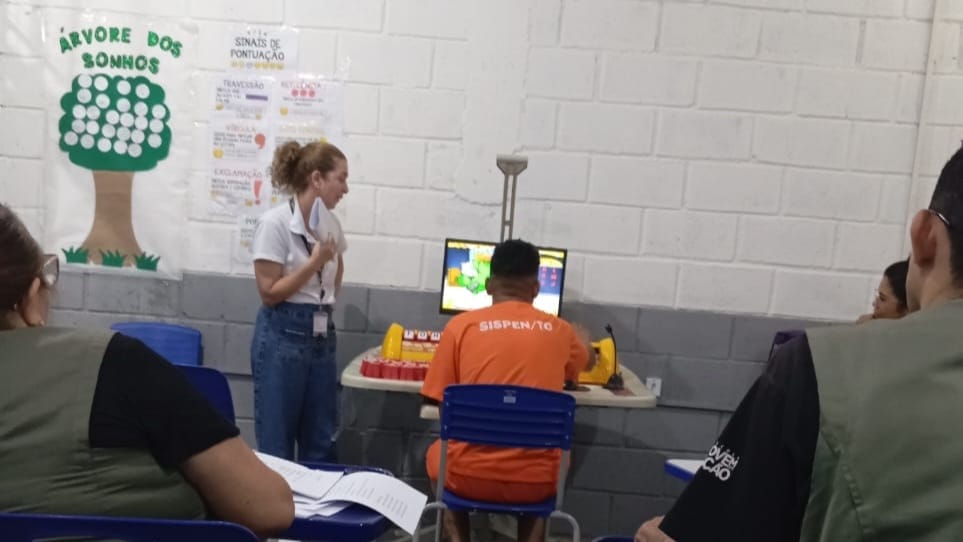 Homem negro, presidiário, sentado de costas numa cadeira azul, usando uniforme laranja. Ele está usando uma mesa educacional, recurso que ajuda na alfabetização, ao lado dele, em pé, uma instrutora branca de cabelos cacheados e presos, usa calça jeans e camiseta branca. A mesa está encostada em uma parede metade cinza, metade branca