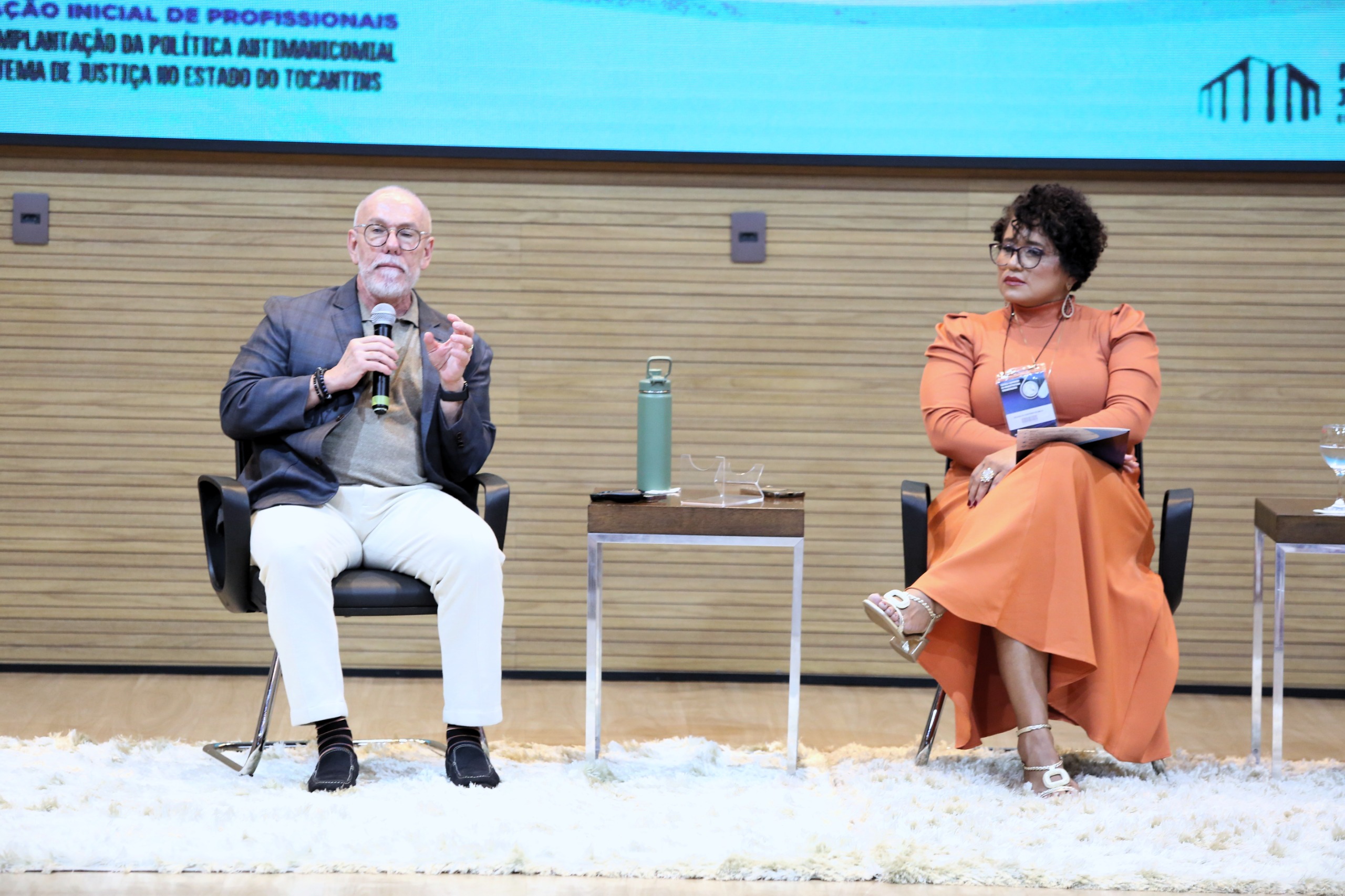 Imagem mostra um telão colorido ao fundo, à frente uma mulher com roupa laranja sentada e um homem branco, calvo com calça branca e terno azul falando ao microfone