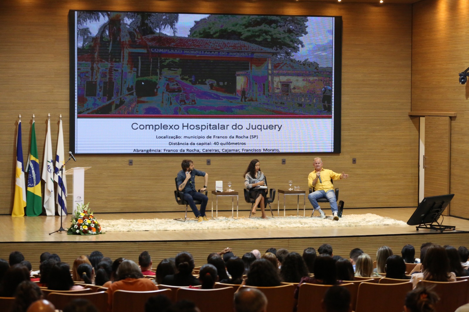 Fotografia colorida que mostra dois homens (um vestindo preto e outro amarelo) e uma mulher morena e jovem ao centro (ela usa calça preta e blusa listrada). Eles estão no palco, sendo  observado por uma plateia. Ao fundo tem um telão com foto do Complexo Hospitalar do Juquery