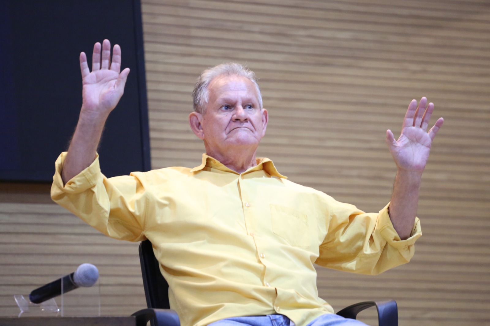 Fotografia colorida que mostra um homem branco, idoso, usando camisa amarela e de braços abertos