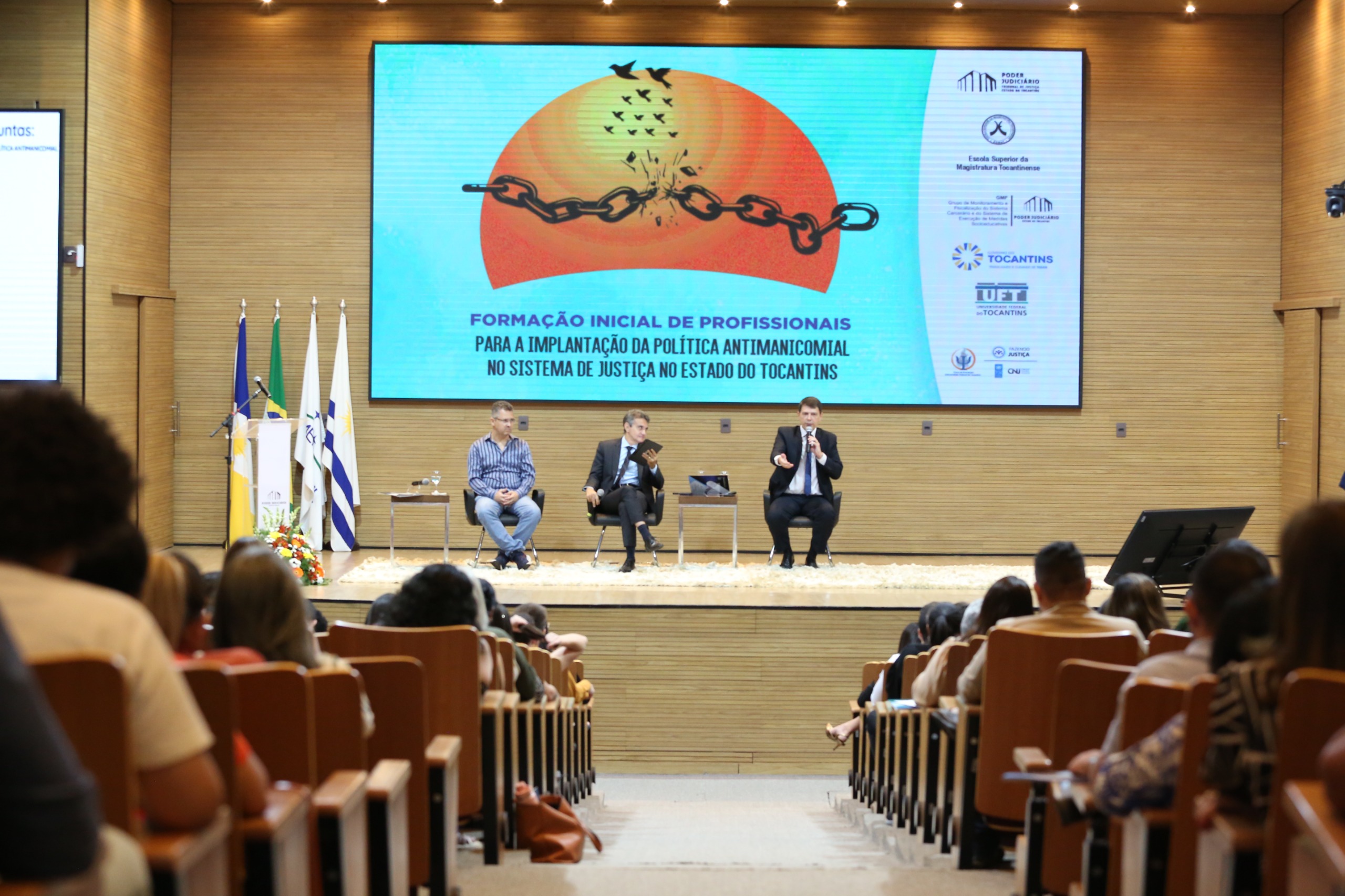 Imagem de um auditório com várias pessoas sentadas e no palco três pessoas sentadas; ao fundo um telão