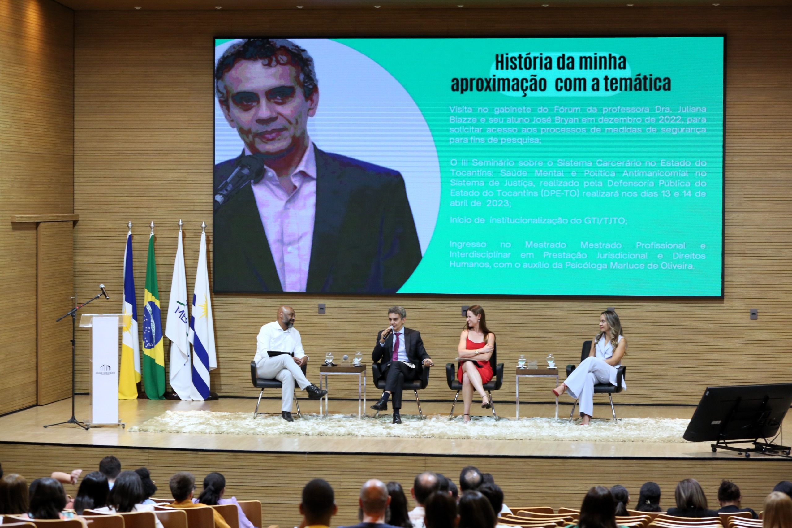 Auditório com várias pessoas sentadas e no palco, bandeiras do Brasil, do Tocantins e pessoas sentadas; ao fundo um telão